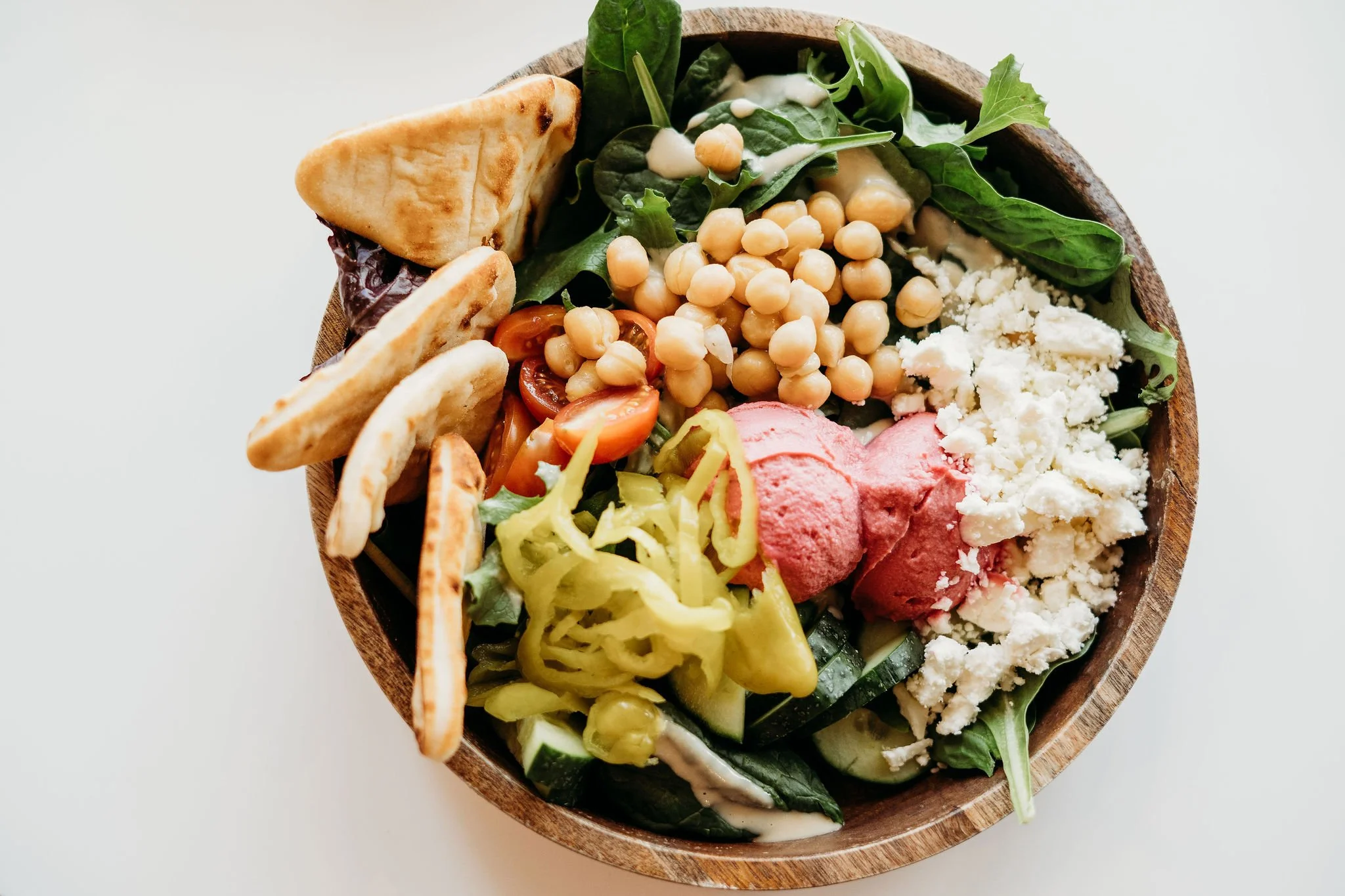 A wooden bowl filled with a colorful vegetable salad, including cherry tomatoes, cucumber, lettuce, spinach, chickpeas, crumbled feta cheese, pink hummus, and pita chips, on a white background.
