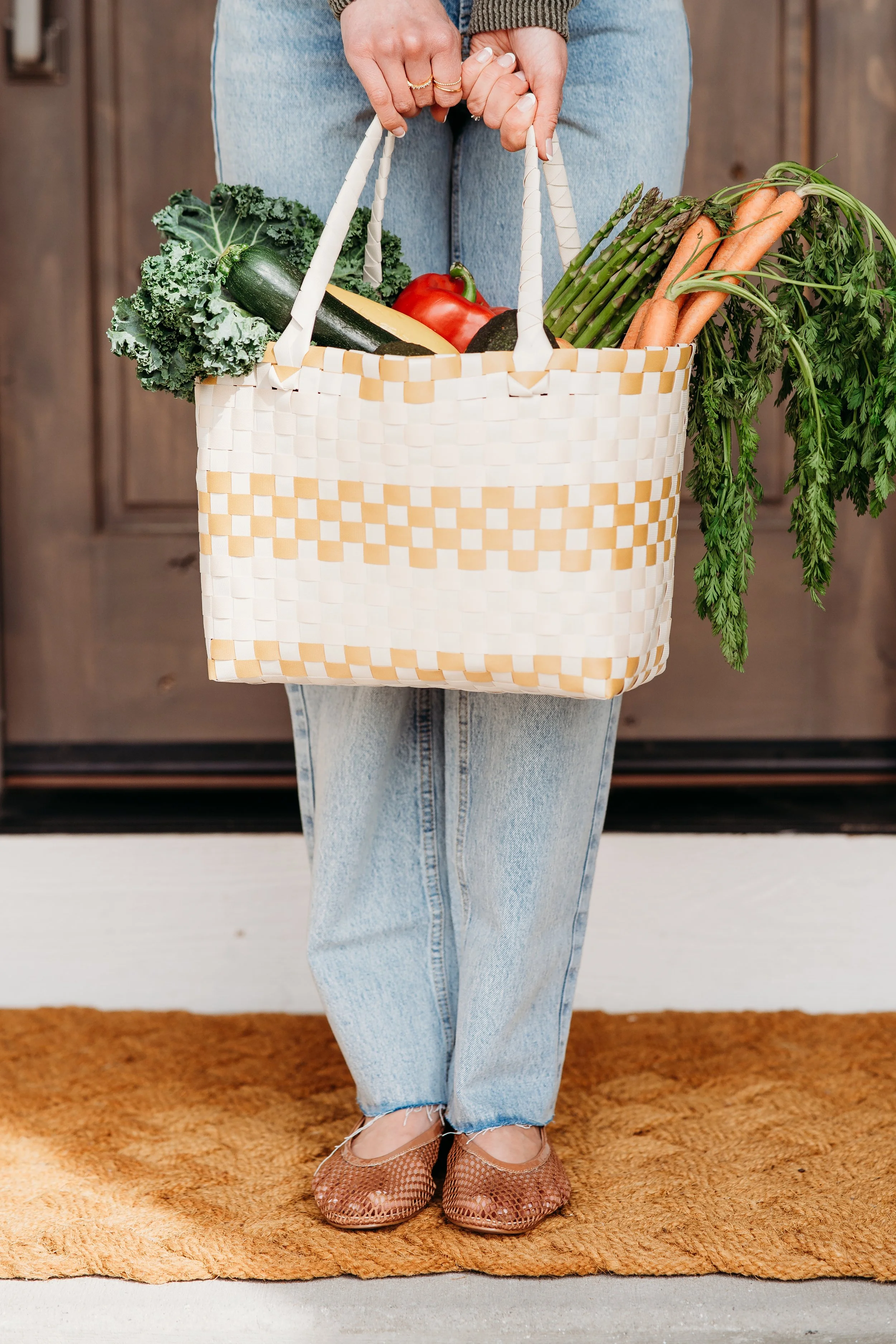 Person standing indoors holding a woven basket filled with vegetables including carrots, zucchini, leafy greens, tomatoes, and asparagus.