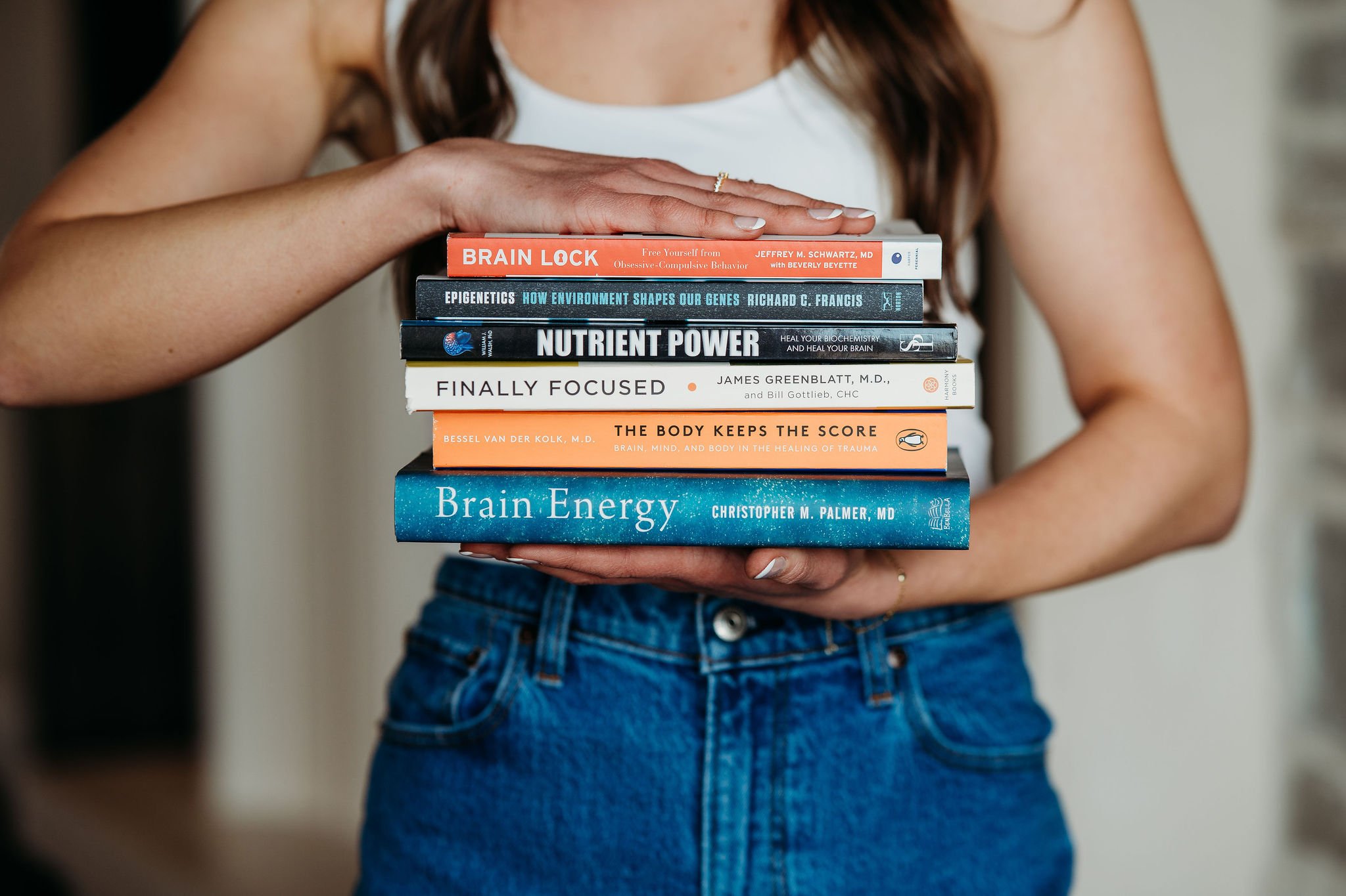 Person holding a stack of seven health and neuroscience books in front of their torso, wearing a white top and blue jeans.
