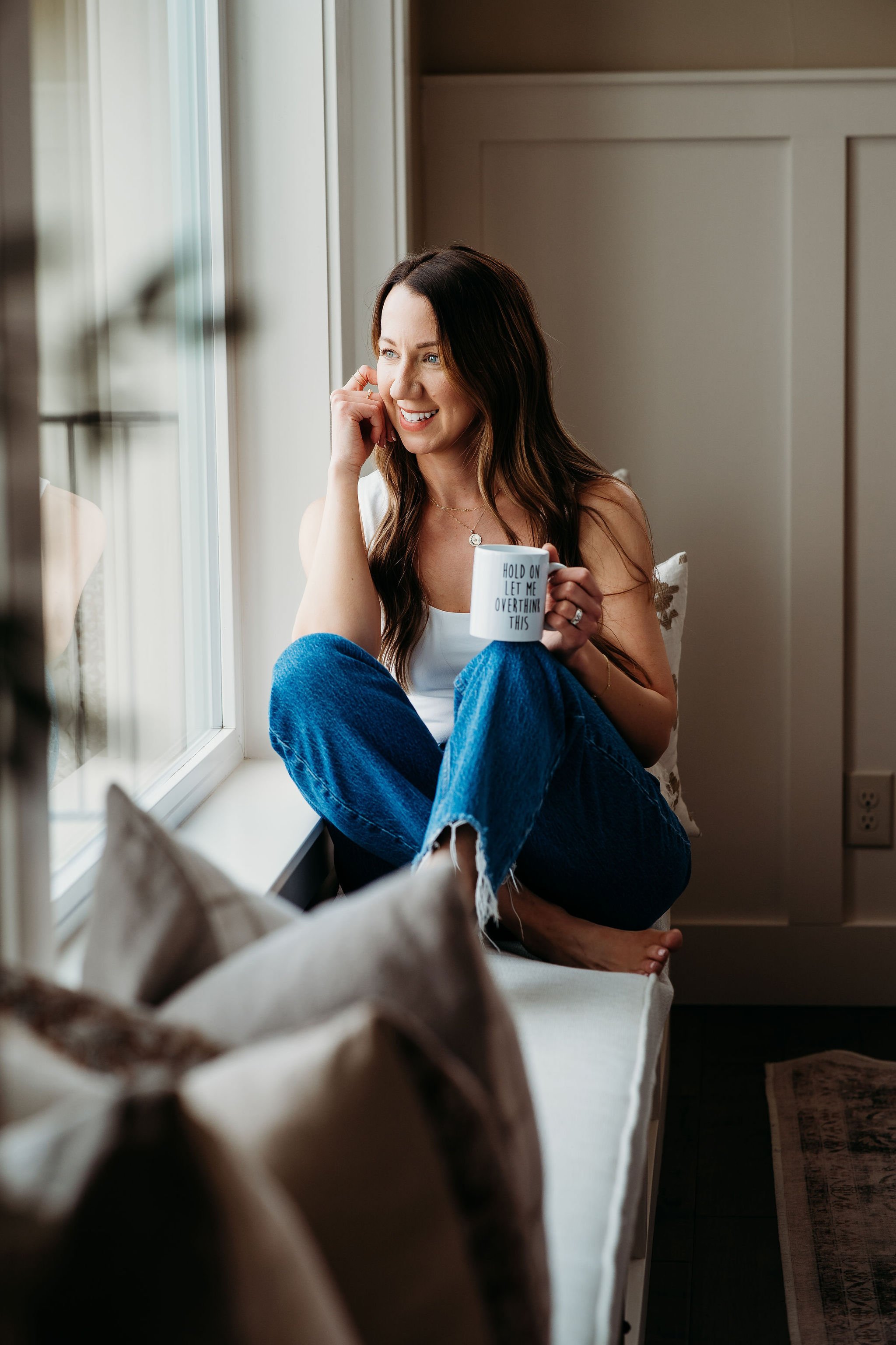 A woman sitting on a window seat, holding a mug with the words 'Hold on let me overthink this' written on it. She is smiling and looking out the window.