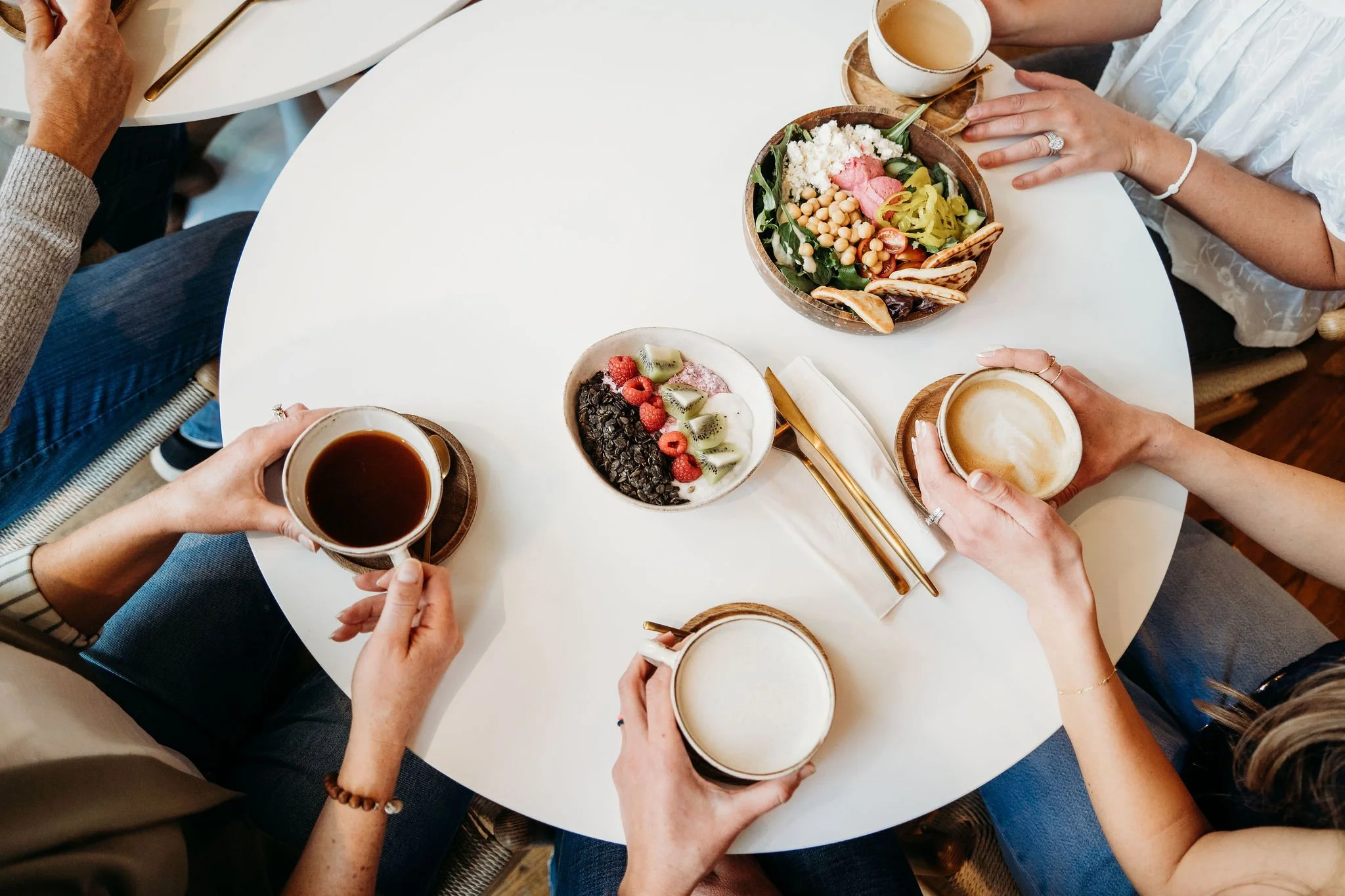 People enjoying coffee, a salad, a yogurt bowl with berries and kiwi, and a cup of tea or coffee at a round white table.