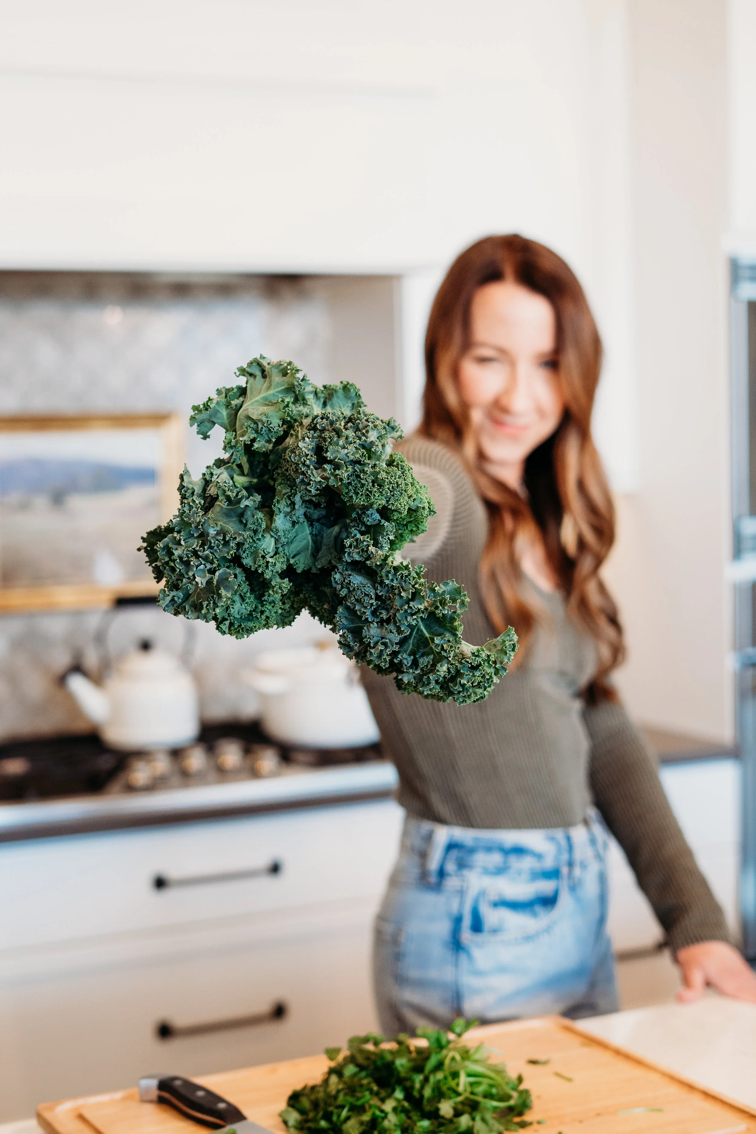 A woman with long, wavy brown hair holding a bunch of kale in a kitchen.