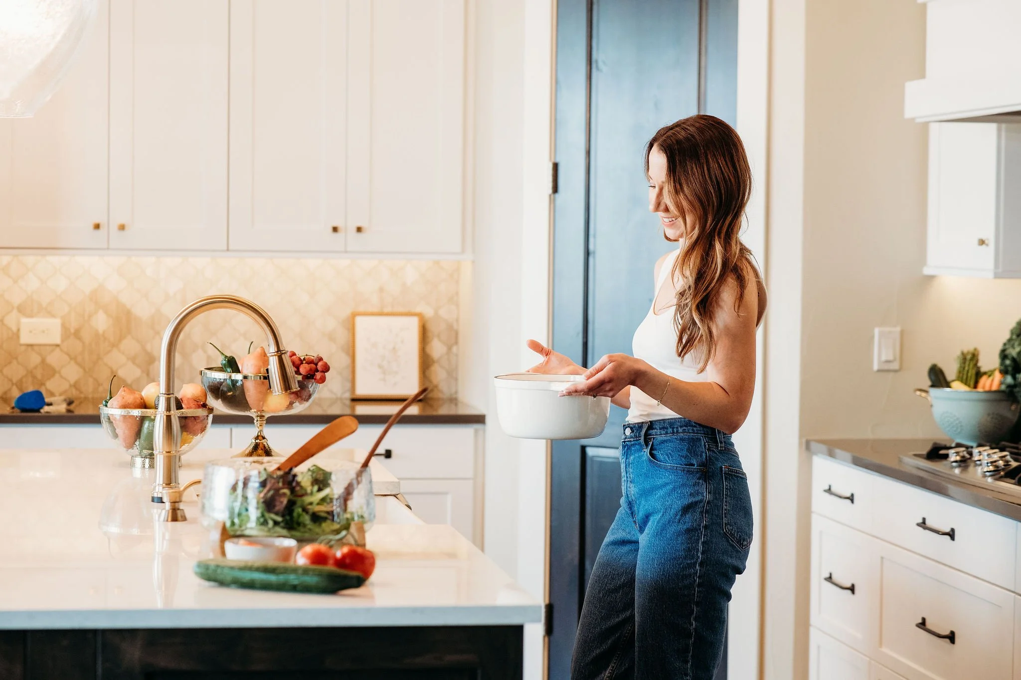 A woman in a white tank top and blue jeans standing in a modern kitchen, smiling and holding a white bowl, with various fruits and vegetables on the kitchen counter.