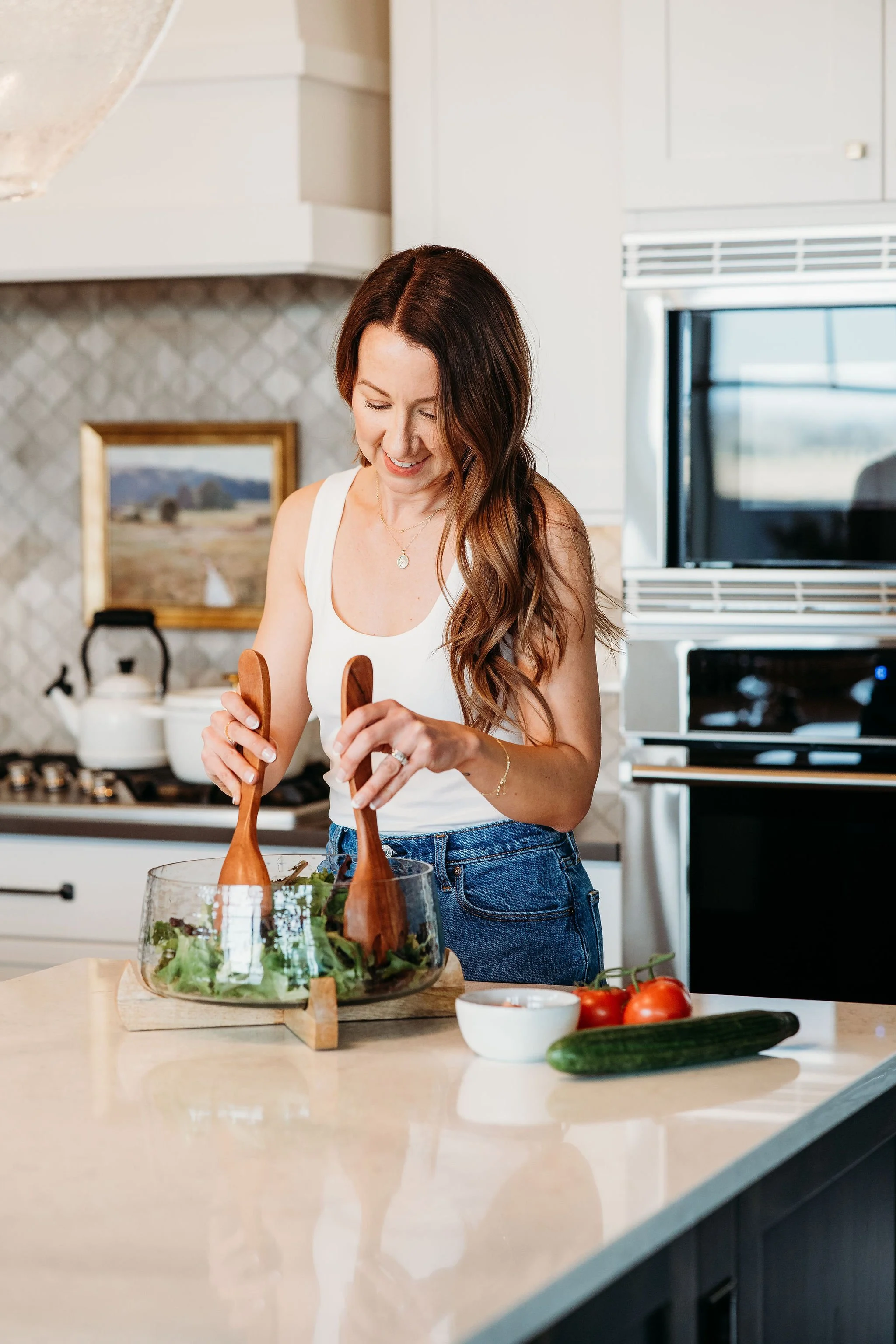 A woman with long wavy brown hair, wearing a white tank top and blue jeans, is smiling while tossing salad in a glass bowl in a bright modern kitchen.