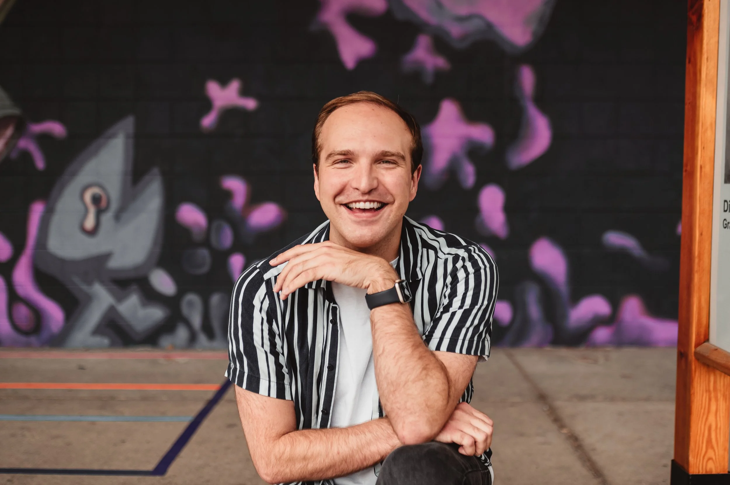 Cameron Stefanski grins into the camera with his arms resting on one raised knee. He wears a black and white striped shirt, a white undershirt, and a smartwatch with a black band.