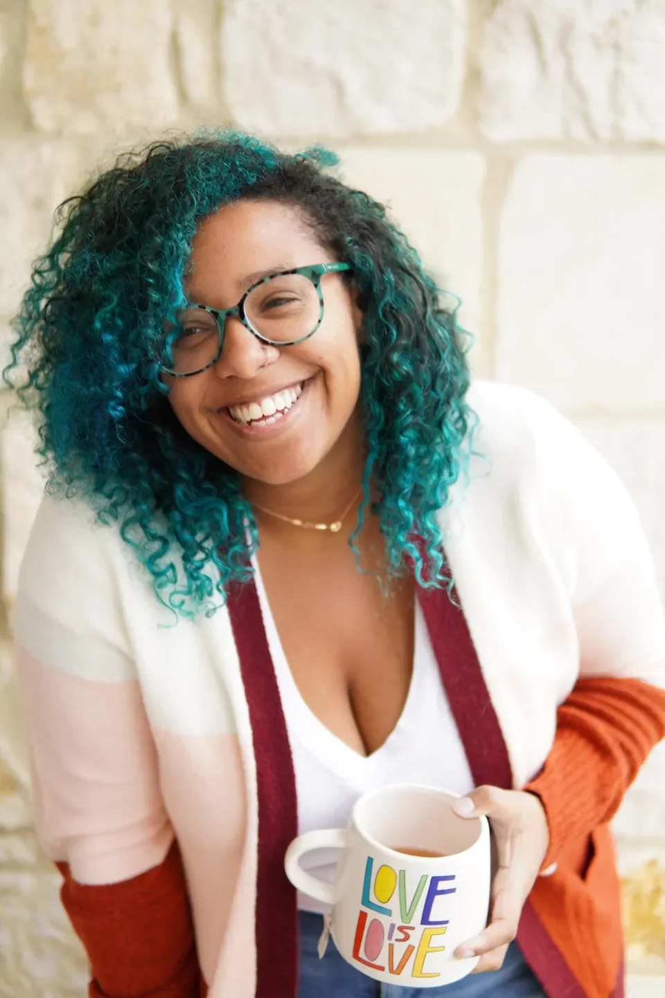 Madison Butler smiles to someone off-camera. Her curly natural hair is dyed a bright and cheerful blue, and she wears a warm multi-toned cardigan. She's holding a mug that says "Love Is Love"