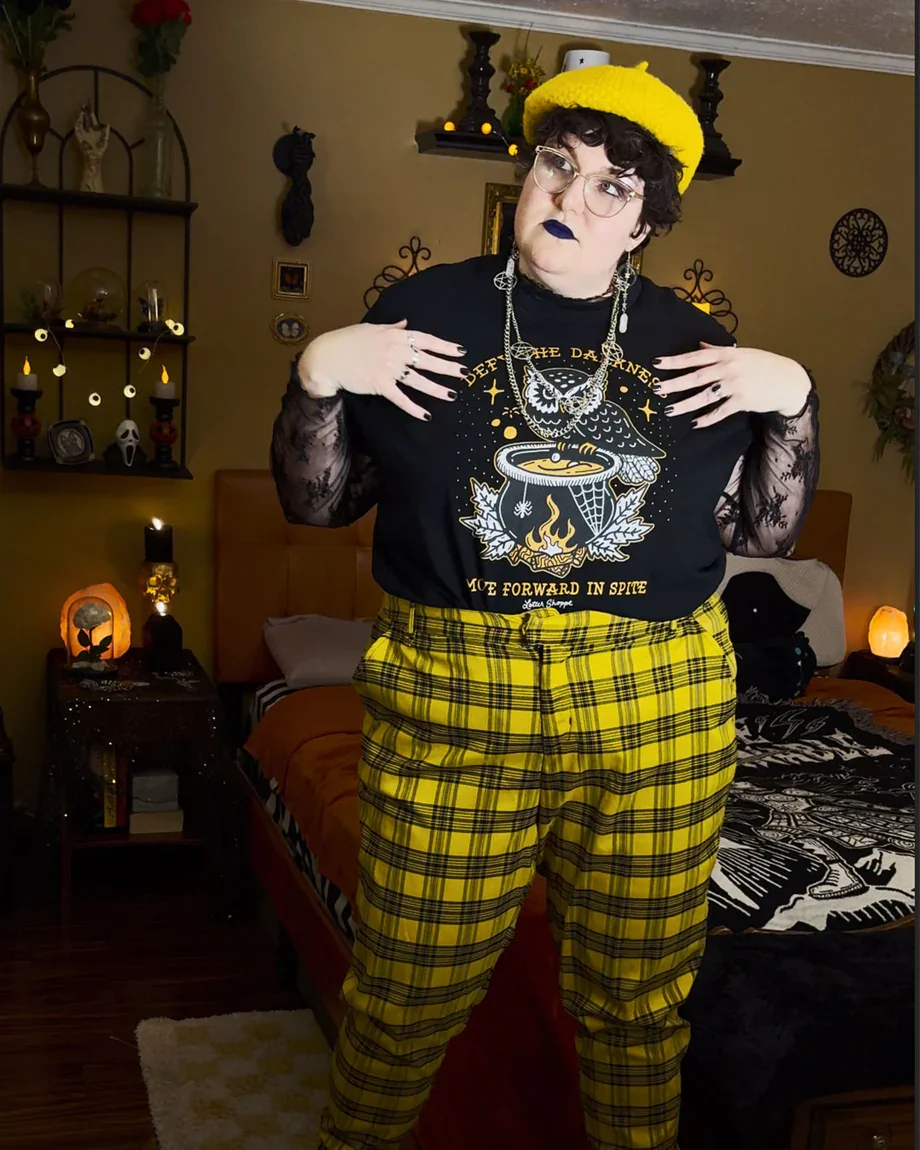 Dean wearing a bright yellow beret, yellow plaid pants, heavy silver jewelry, and black nail polish and lipstick, modeling an outfit from Letter Shoppe with their artwork on it. They gaze to their right, off-camera.