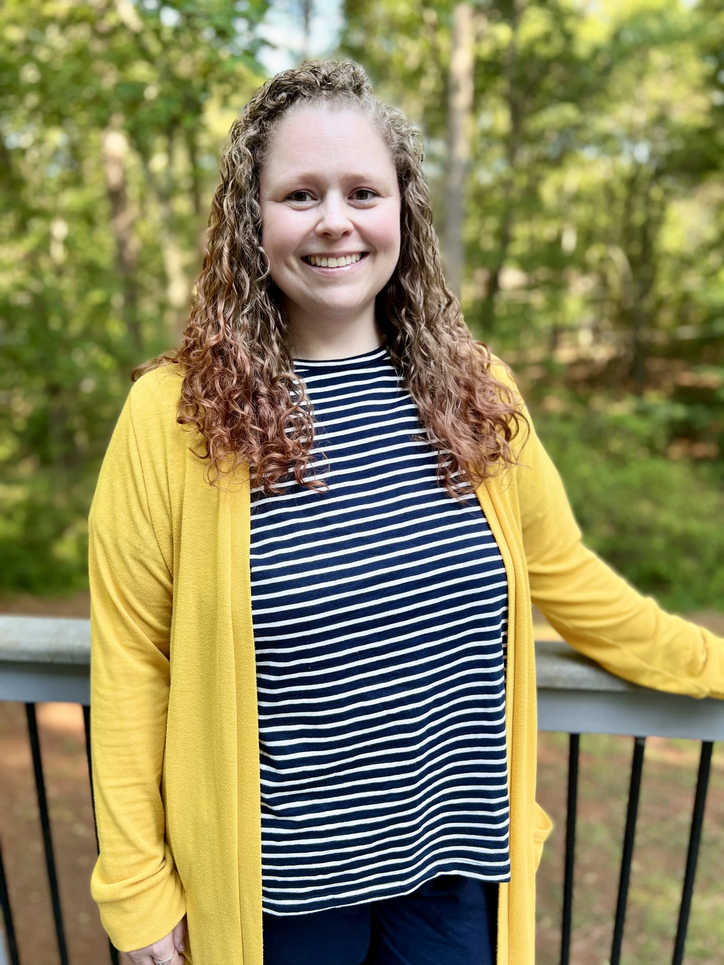 A woman with curly hair wearing a navy and white striped shirt and a yellow cardigan, standing outdoors in front of a green wooded background and a park railing.