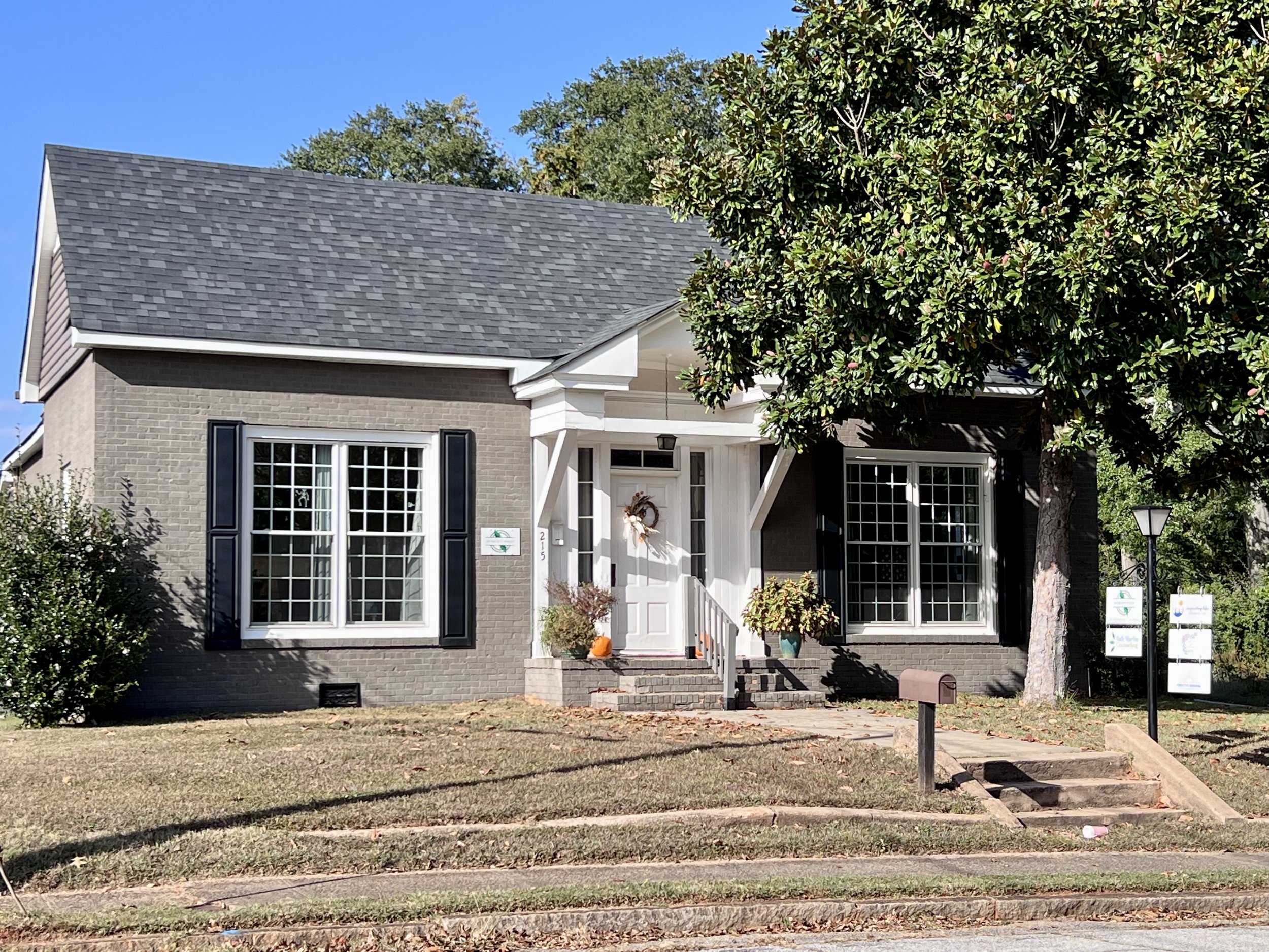 Front view of a house with grey brick exterior, white door, large windows, and black shutters. There is a small front porch with potted plants, and a mature tree partially shading the house. The lawn is trimmed, and steps lead to the sidewalk.
