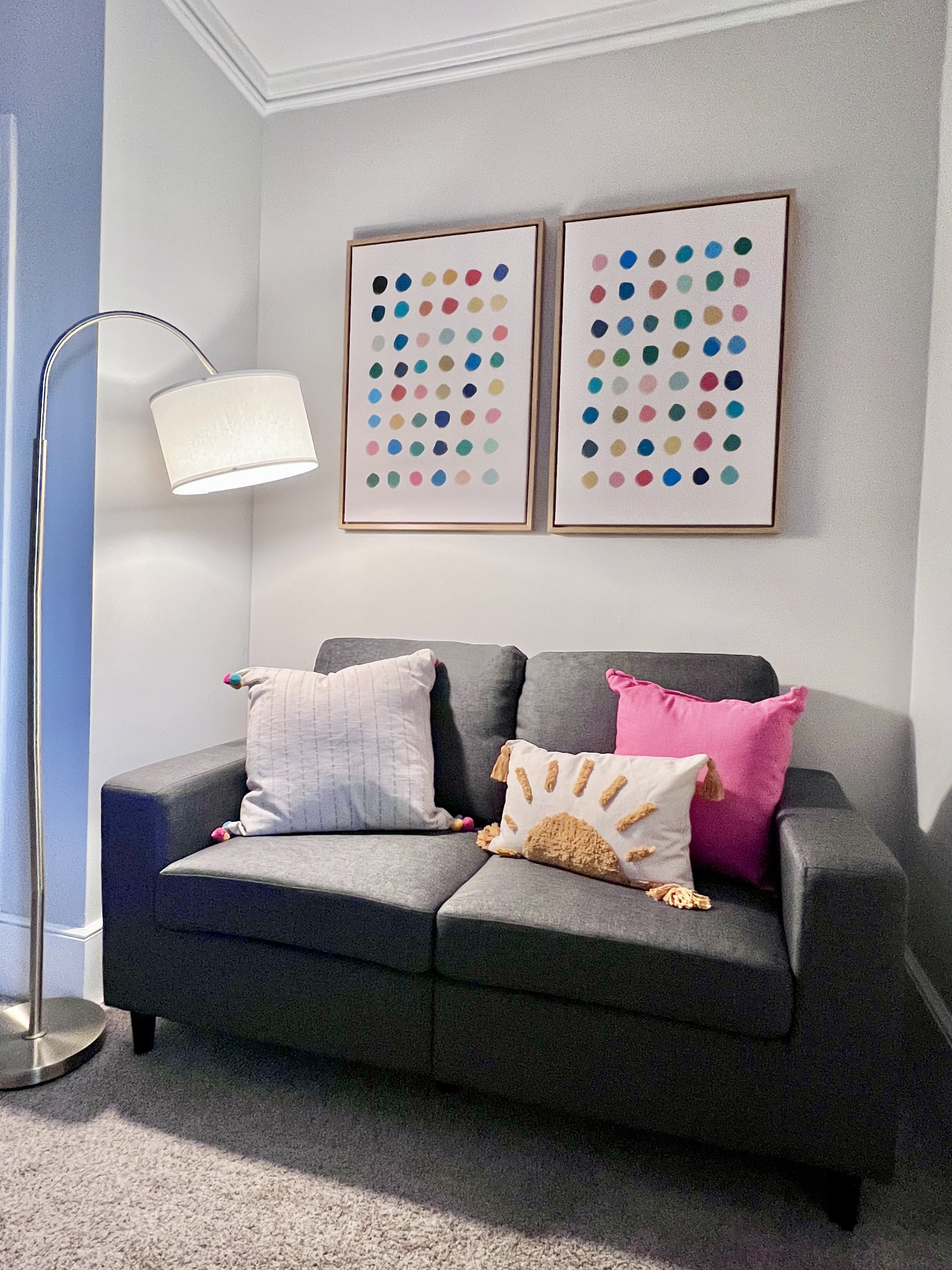 A living room with a dark gray couch, three colorful pillows, and two framed art prints above, and a tall floor lamp with a white shade.