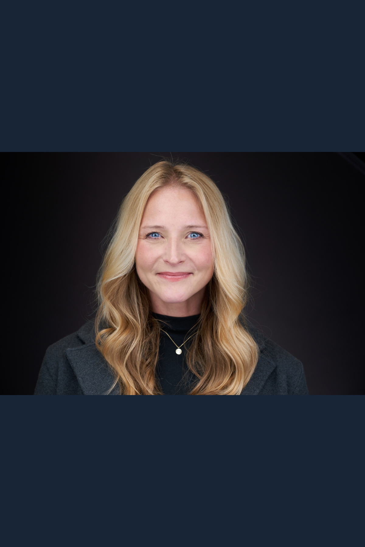 A portrait of a woman with long, wavy blonde hair, blue eyes, and a friendly smile, wearing a dark blazer and a black turtleneck with a pearl necklace, against a dark background.