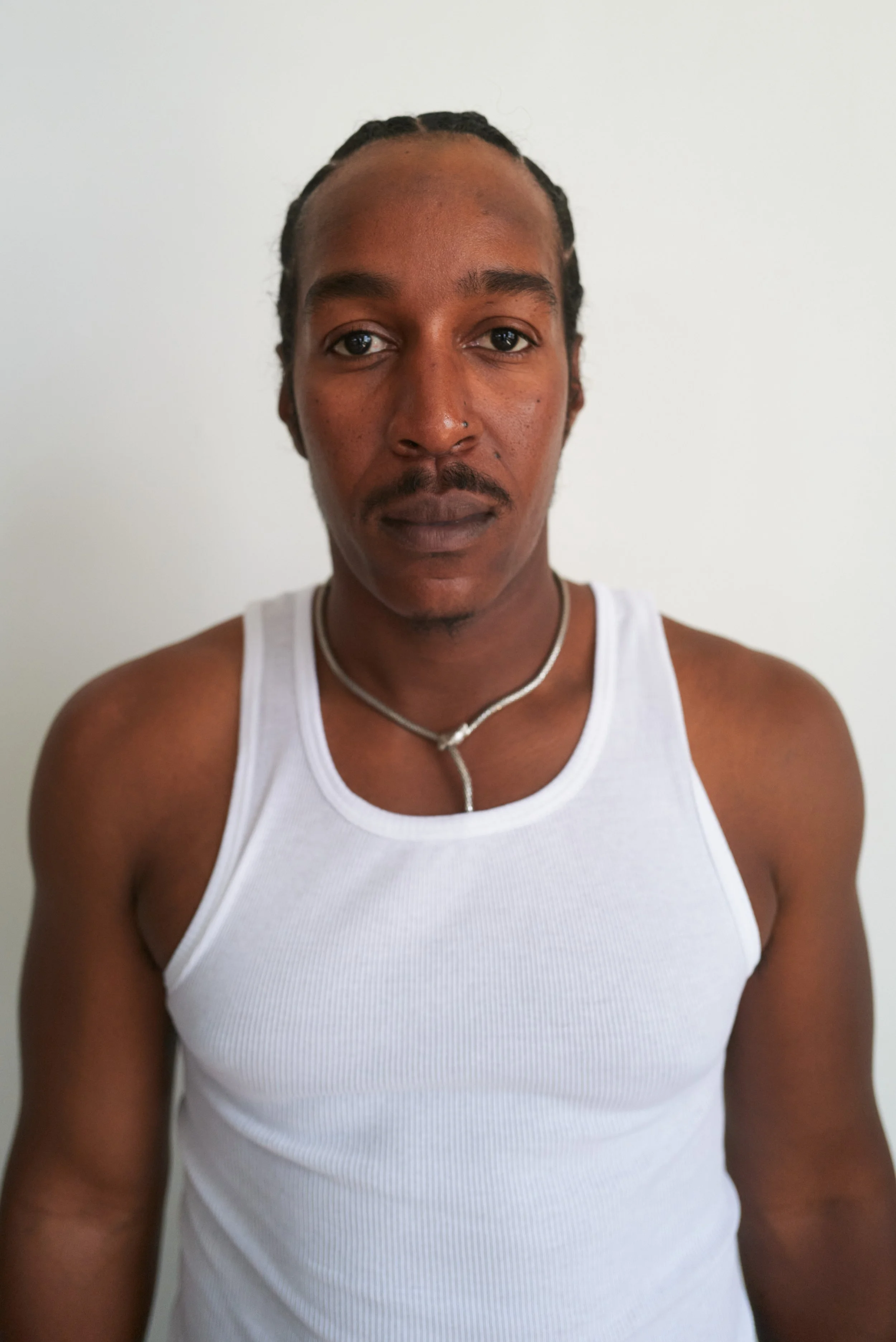 A man with braids, wearing a white tank top and a silver necklace, standing against a plain white background.