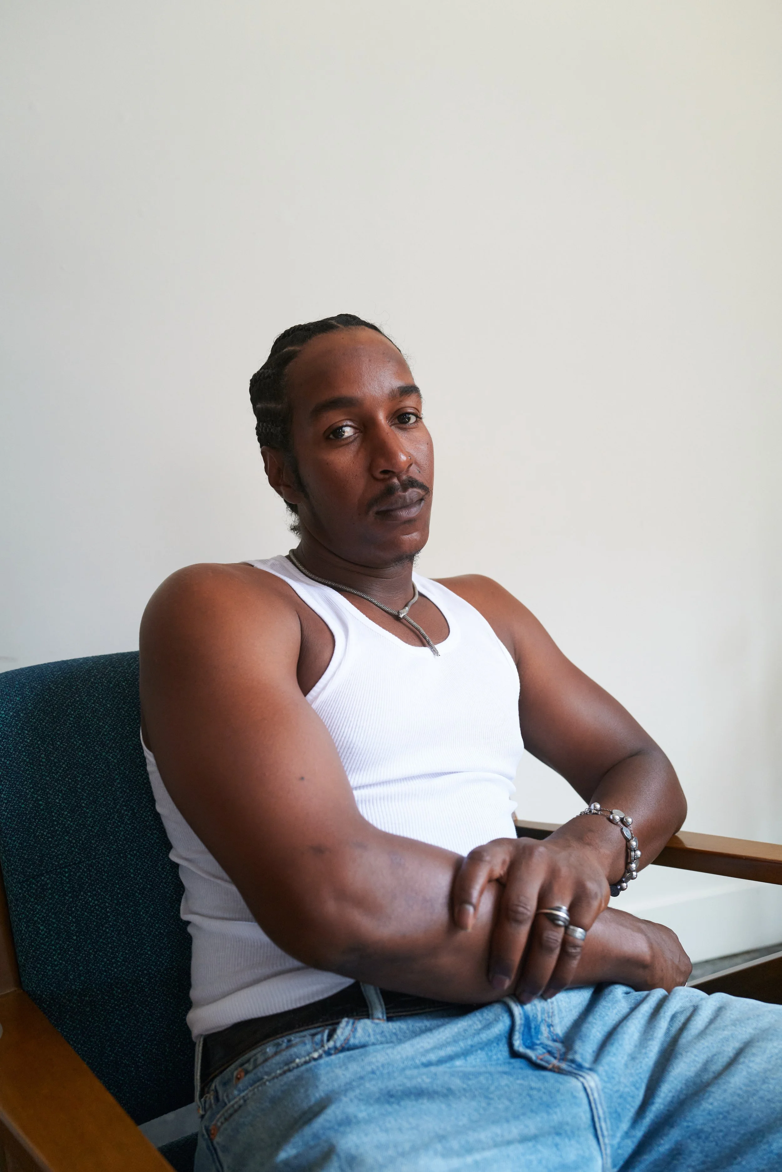 A man wearing a white tank top and blue jeans sitting on a chair with his arms crossed, looking at the camera.