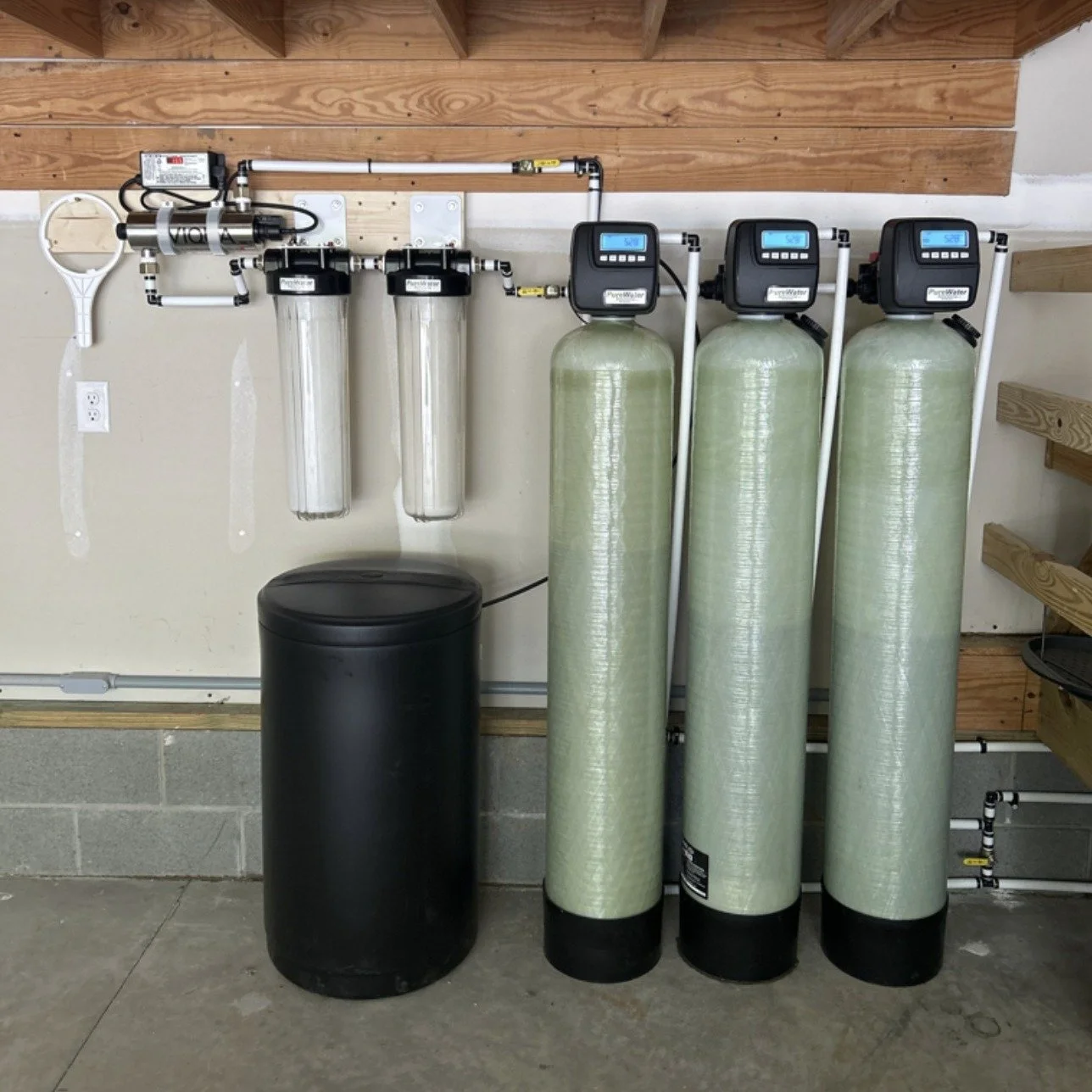 Water filtration system with a digital control valve, large green filter tank, blue casing filter, and black storage tank on a gray speckled floor.