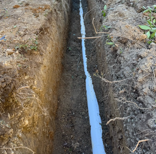 A narrow trench in the soil with a white pipe laid inside.