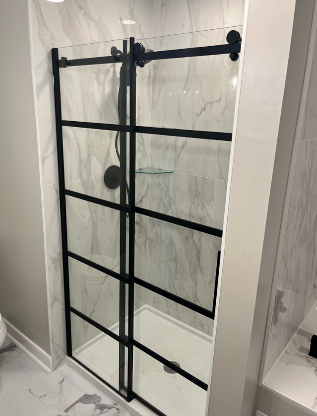 A glass shower door with black metal frame and sliding mechanism in a marble-tile bathroom.