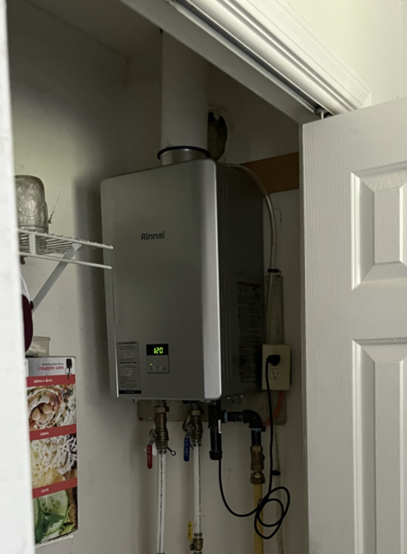 A Rinnai tankless water heater installed in a laundry closet with pipes, valves, and a power outlet.