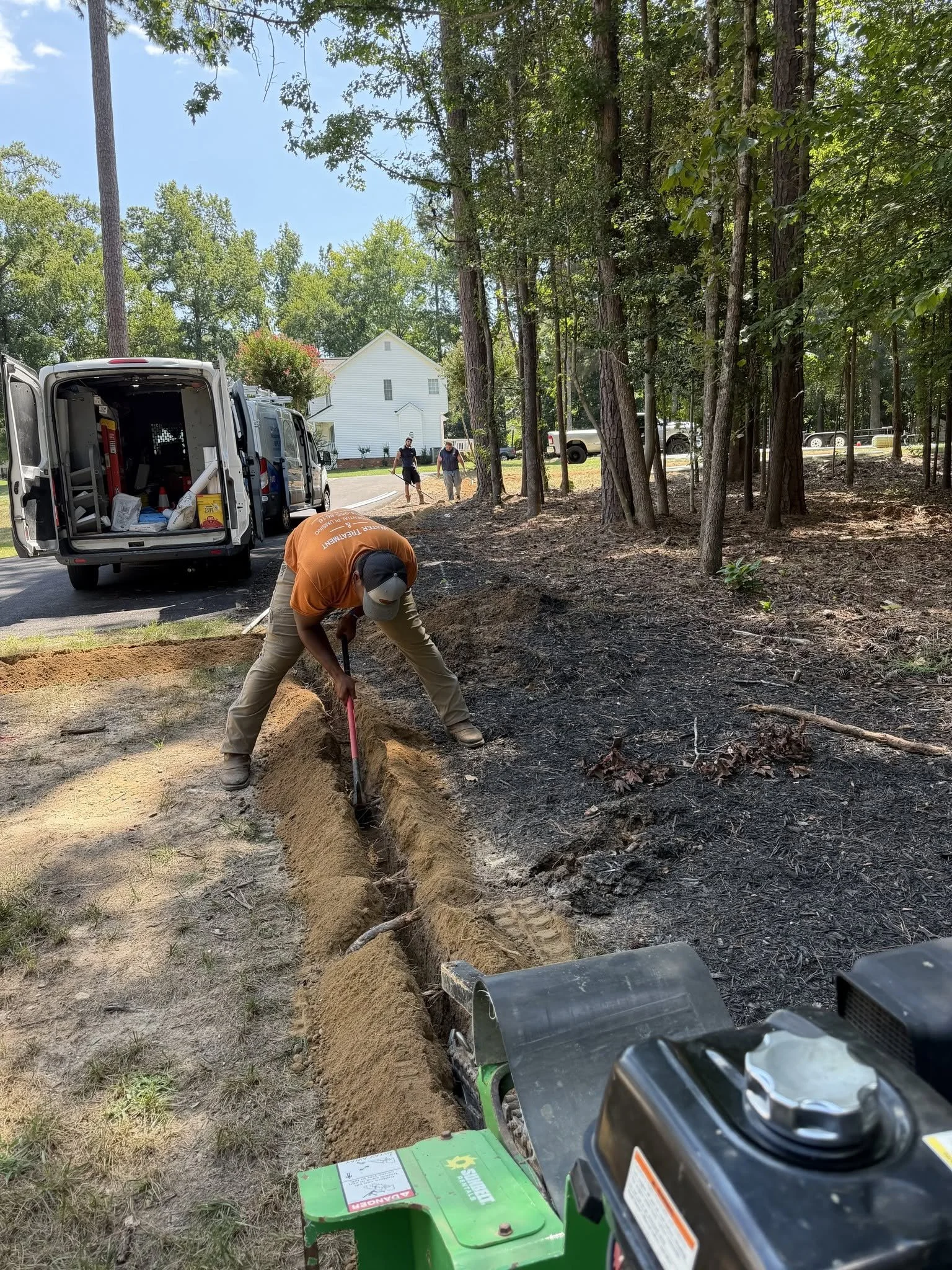 A person wearing an orange shirt and gray cap is working with a shovel in a trench beside a dirt pathway. There are two vehicles parked nearby, with an open utility van filled with supplies. In the background, two people walk along a curving road near a white house, surrounded by tall trees with green leaves and a partly cloudy sky.