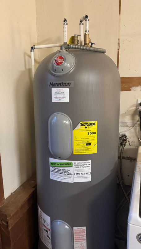 Gray hot water heater in a residential utility room with labels and pipes.
