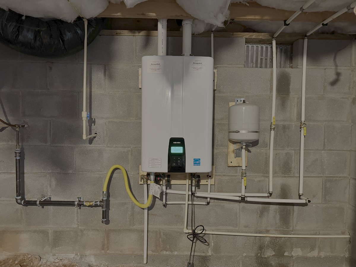 A residential or commercial basement wall with an installed tankless water heater, various pipes, and a pressure expansion tank.