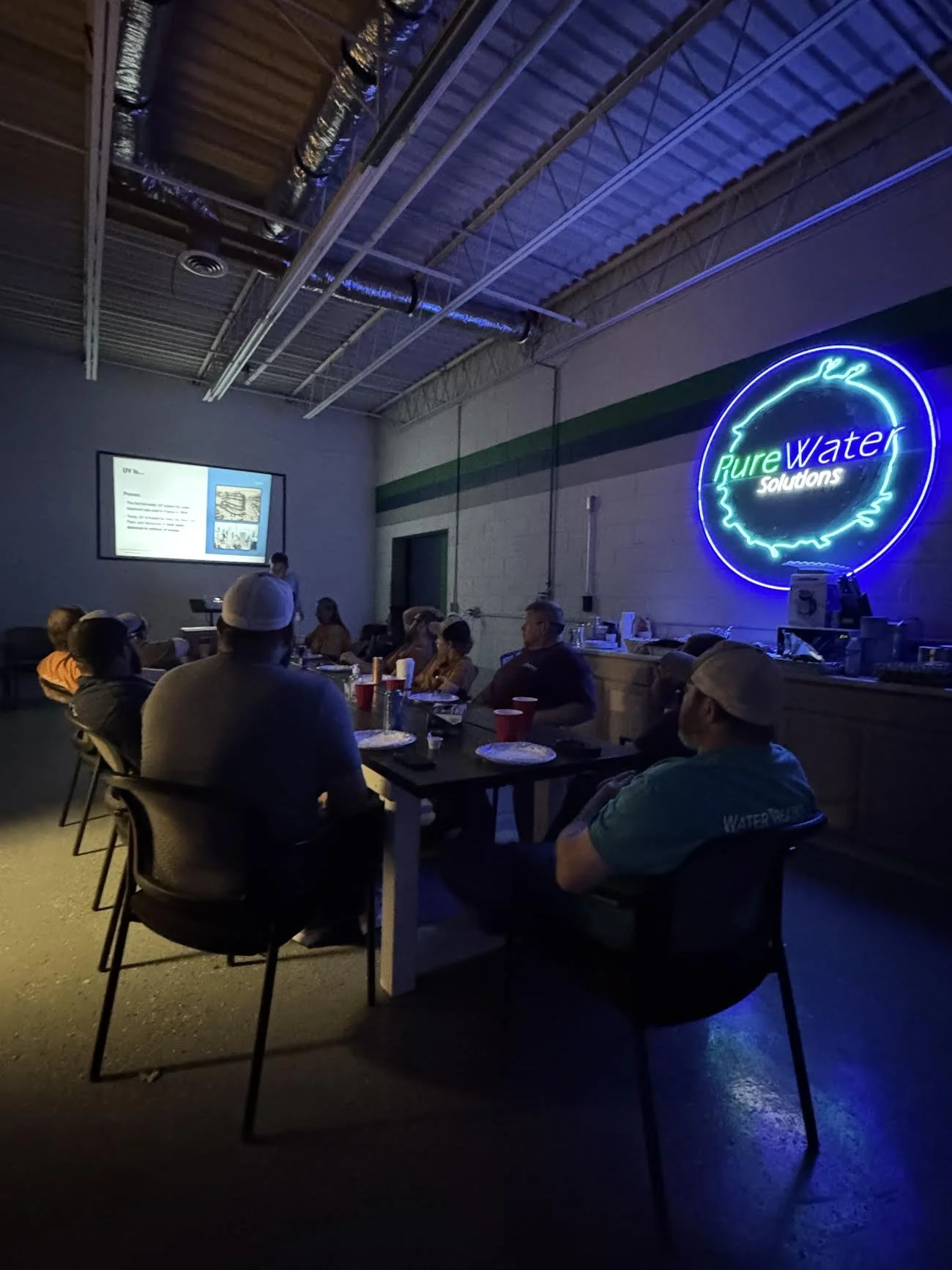 Indoor gathering with people seated at tables watching a presentation, illuminated by a neon sign that reads 'Pure Water Solutions' on the wall.