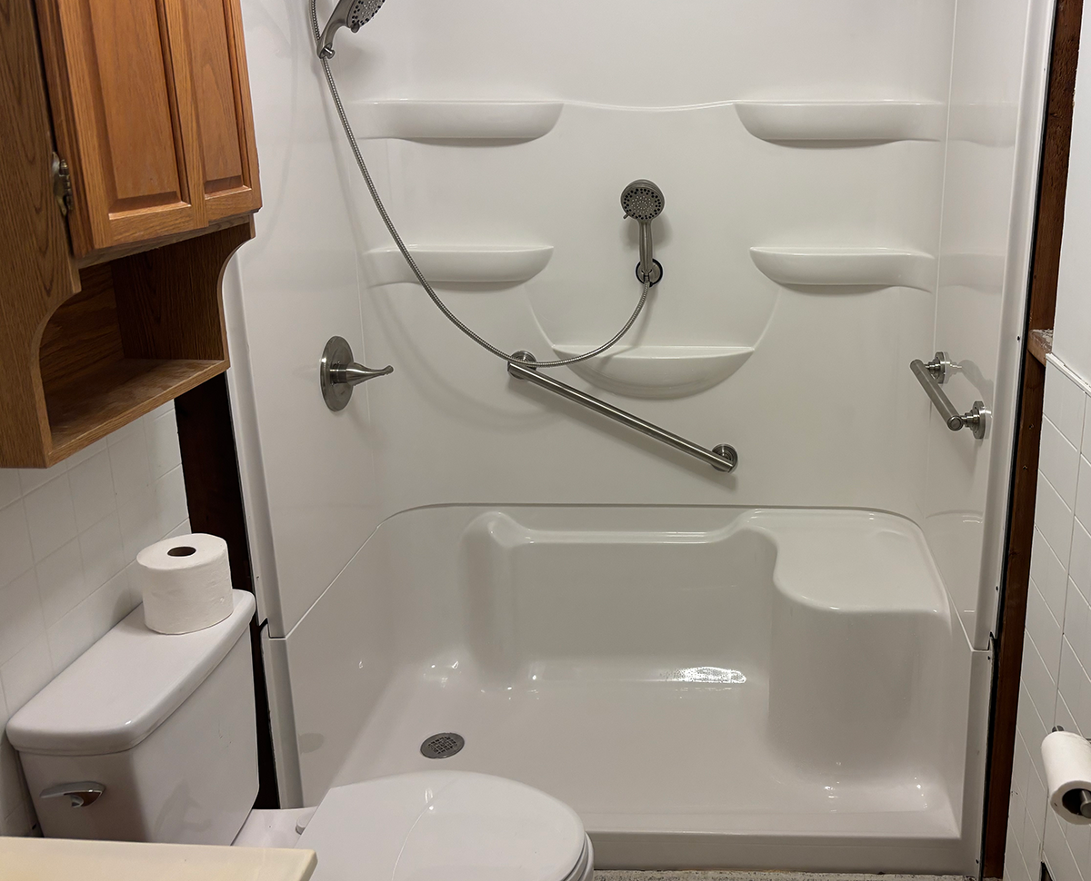 Bathroom with a shower stall featuring a grab bar, shelves, and a handheld showerhead, toilet with a roll of toilet paper, and wooden cabinetry.