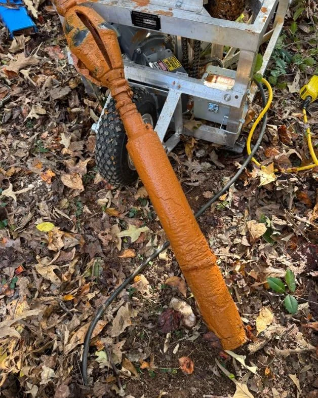 A construction or outdoor tool with a long, rusted, orange-painted metal rod attached to a small metallic machine on the ground surrounded by fallen leaves.