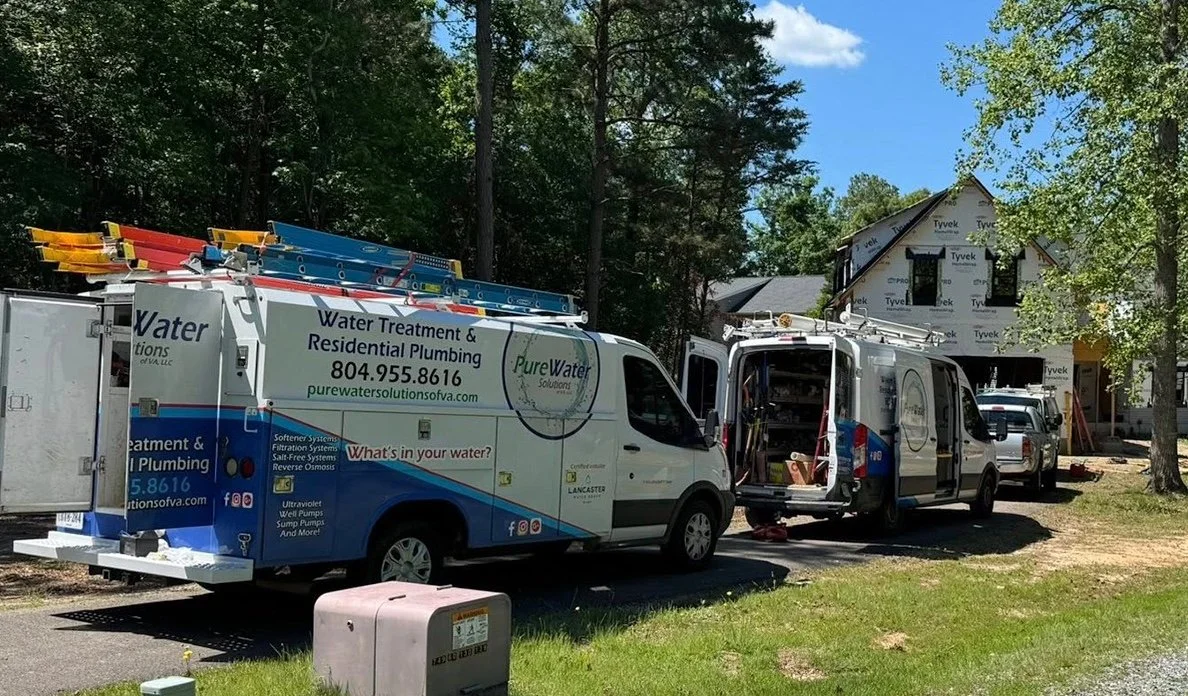 Water solutions company trucks parked at a residential construction site with a house being built. One truck is open, showing tools inside. The trucks display company information and services related to water treatment and plumbing.