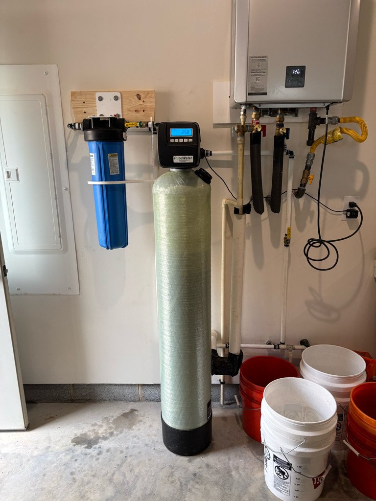 A water filtration system with a blue filter housing, a digital control panel, a tall cylindrical filter, and various pipes and gauges installed on a garage wall, with several empty buckets on the floor.