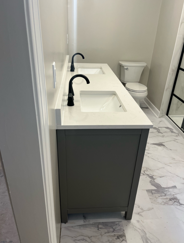 Bathroom with dual sinks, black faucets, gray vanity, and a toilet in the background, marble-patterned floor tiles, and a glass shower door.
