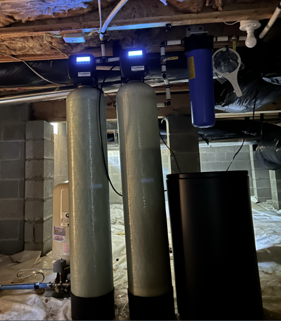 Water filtration system with two large vertical tanks, a blue sediment filter, and water treatment control units installed in a basement with exposed ceiling and concrete block walls.