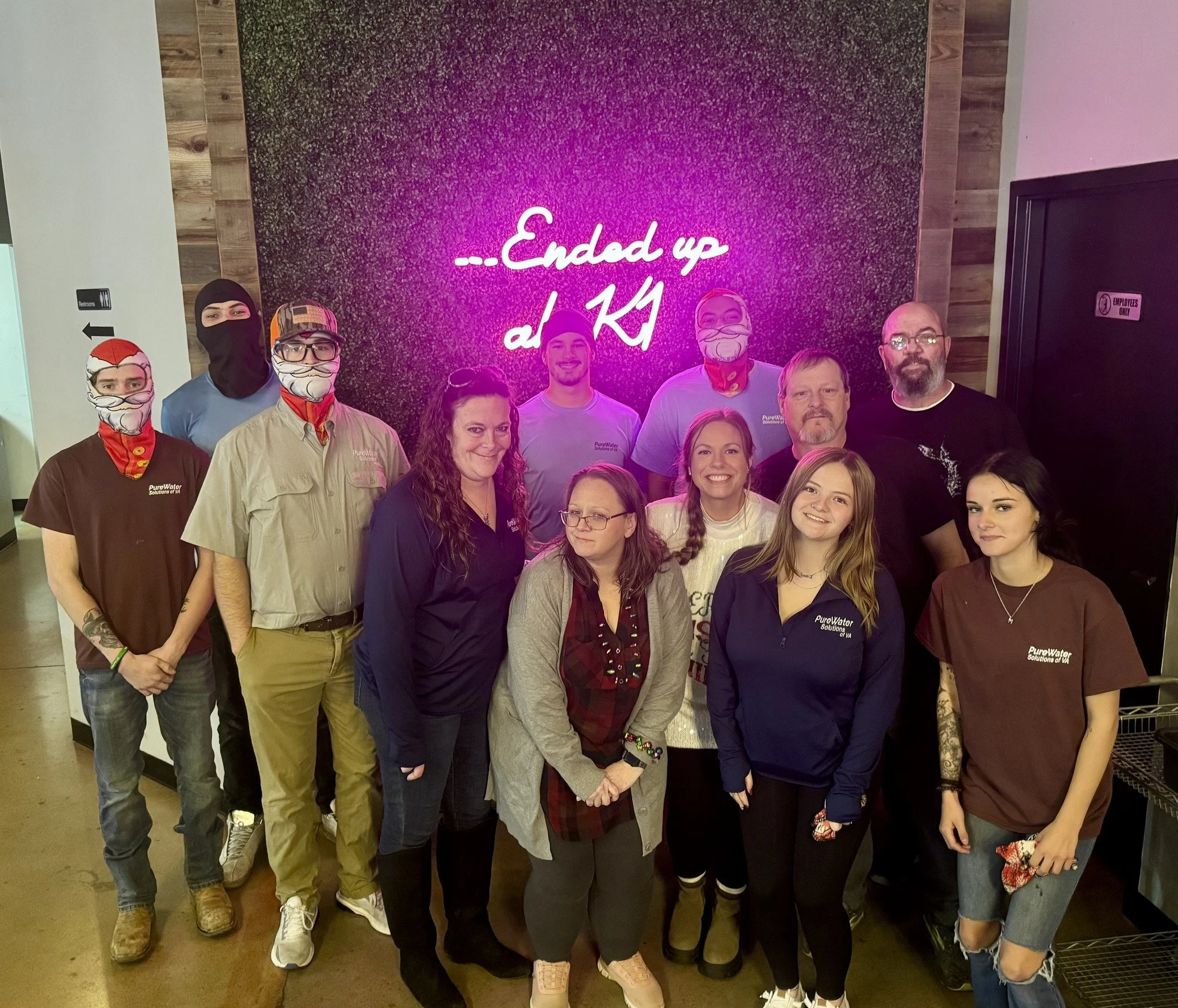 Group of people standing in front of a black wall with pink neon sign that reads 'Endeed up at KI'. Some individuals are wearing masks and uniforms with 'Pure Water.'