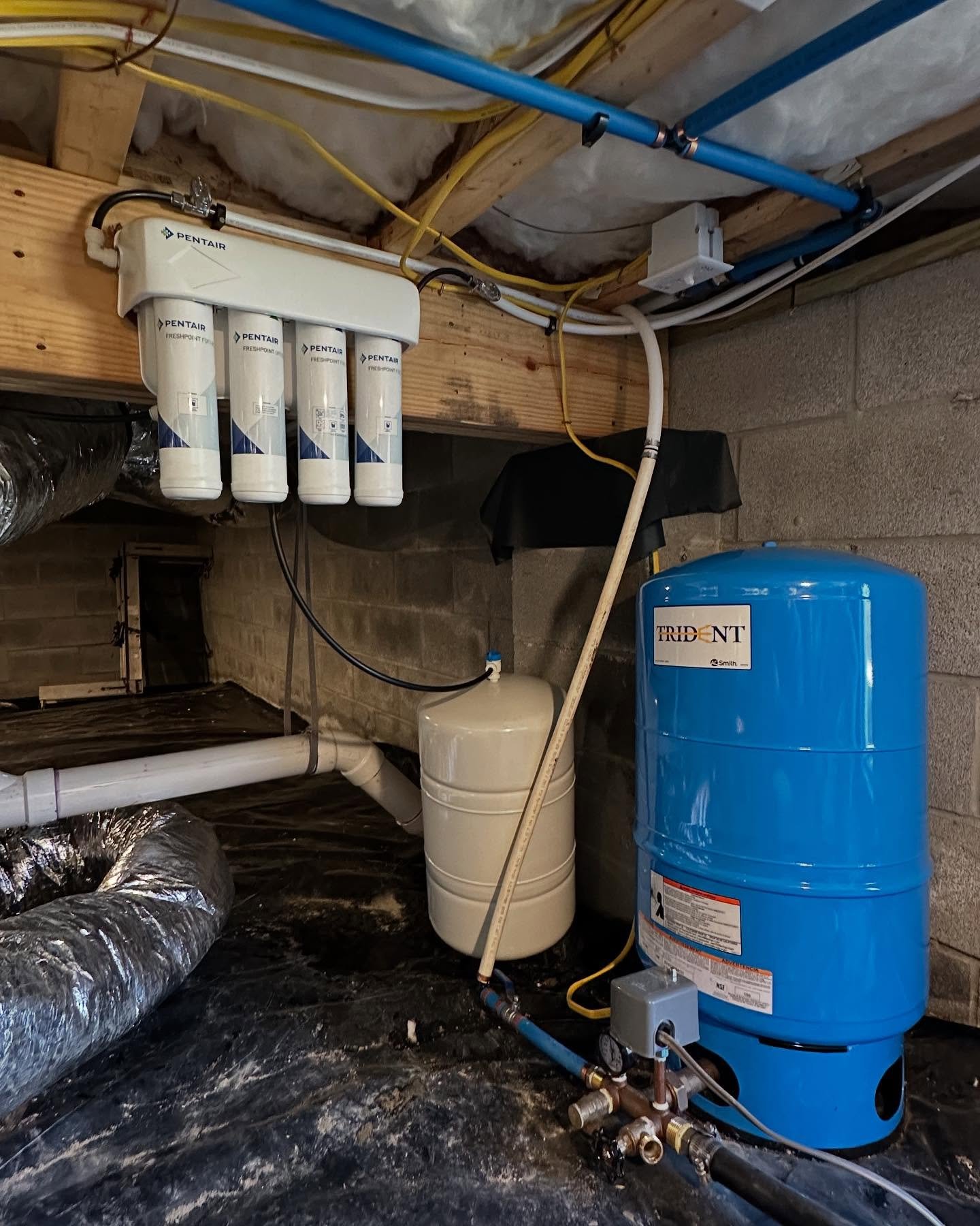 A basement with water filtration system, large orange water tank, and pipes. The filtration unit has four filters labeled PENTAIR, mounted on a wooden beam.