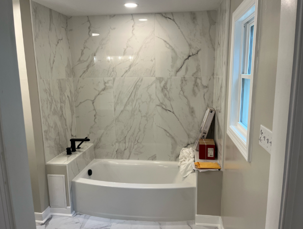 A bathroom with a marble wall and floor, a white bathtub, a window, and some items on a ledge next to the tub.