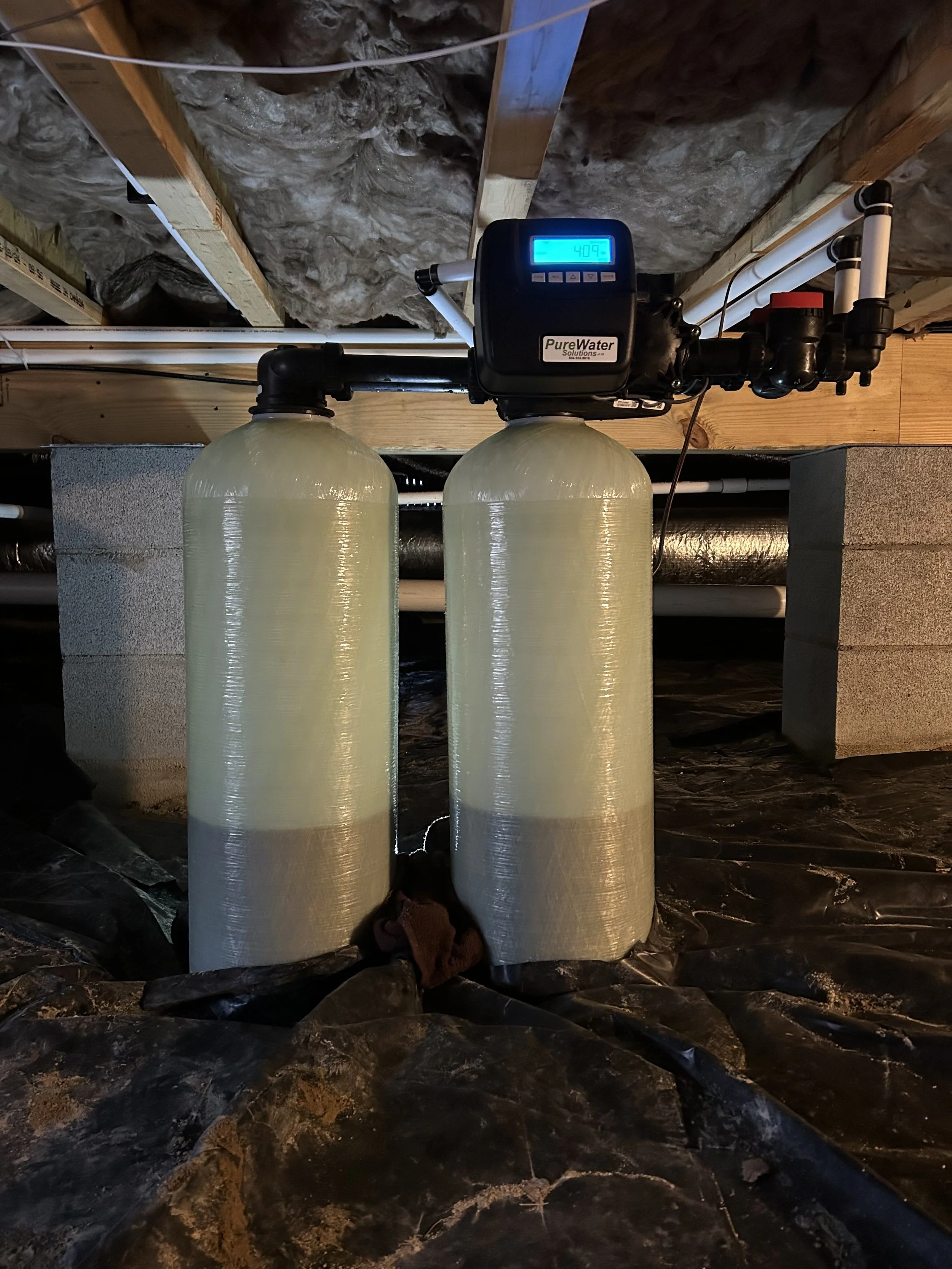 Two large water filtration or softening tanks with a digital control system on top, installed in a basement or utility room, with rooms and construction materials visible in the background.