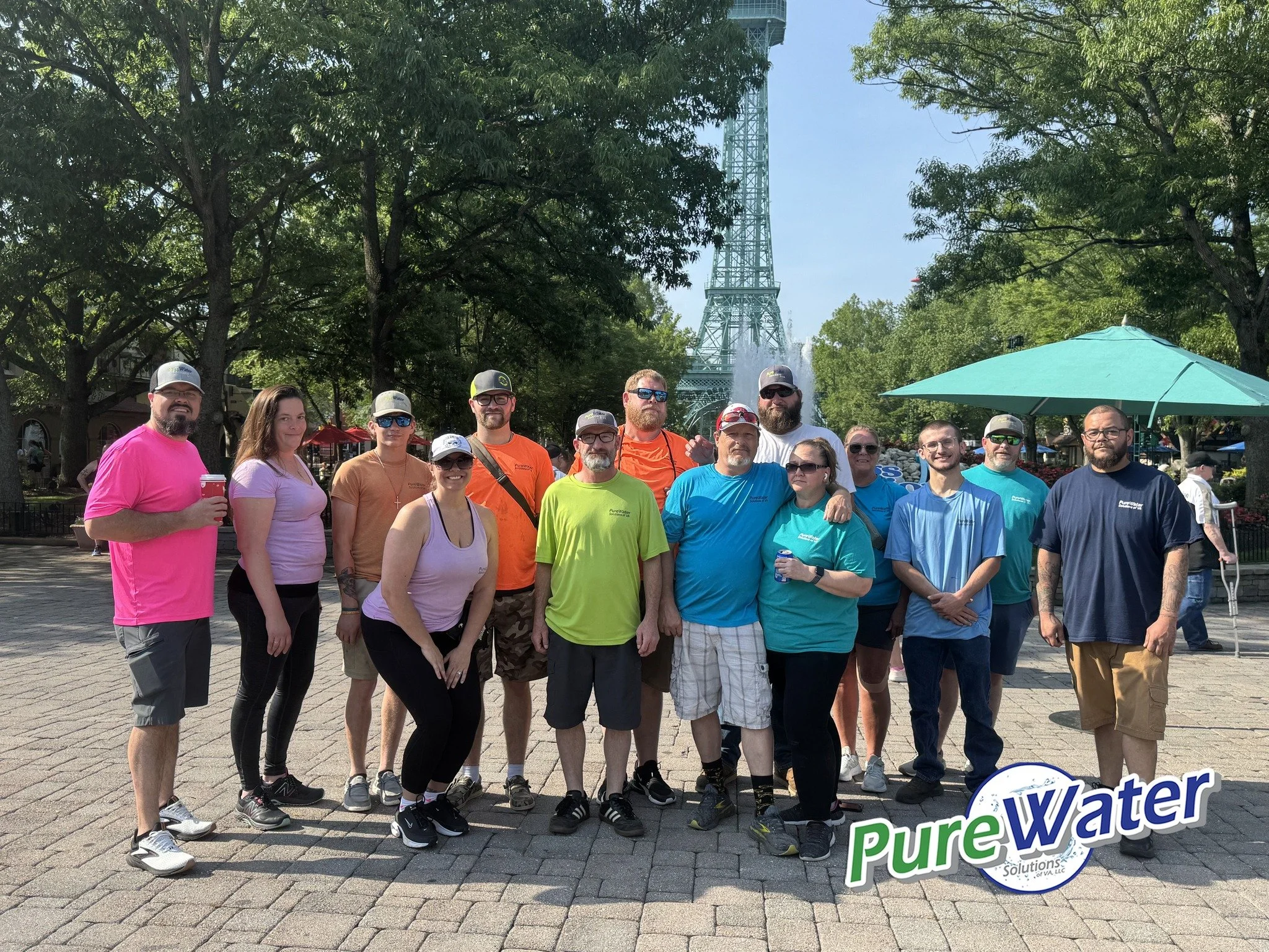 A group of people standing outdoors on a brick path, with trees and the Eiffel Tower in the background. They are smiling, some holding drinks, and wearing casual or athletic clothing. There is a logo for "Pure Water Solutions" in the lower right corner.