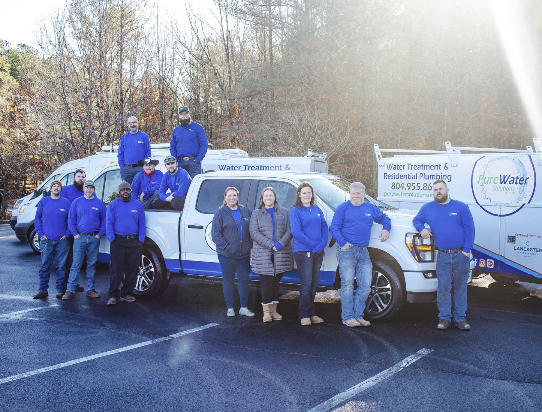 Group of water treatment and plumbing professionals posing in front of company vehicles on a parking lot with trees in the background.