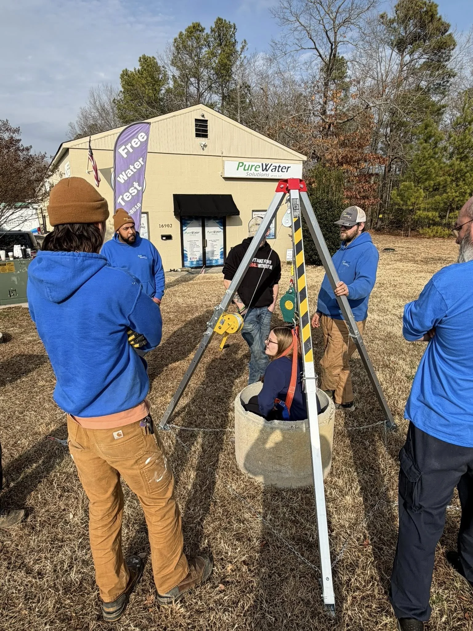 A woman sitting inside a concrete well during a water testing demonstration, surrounded by five men outside a building with a "Pure Water" sign. The building has a banner advertising free water testing.