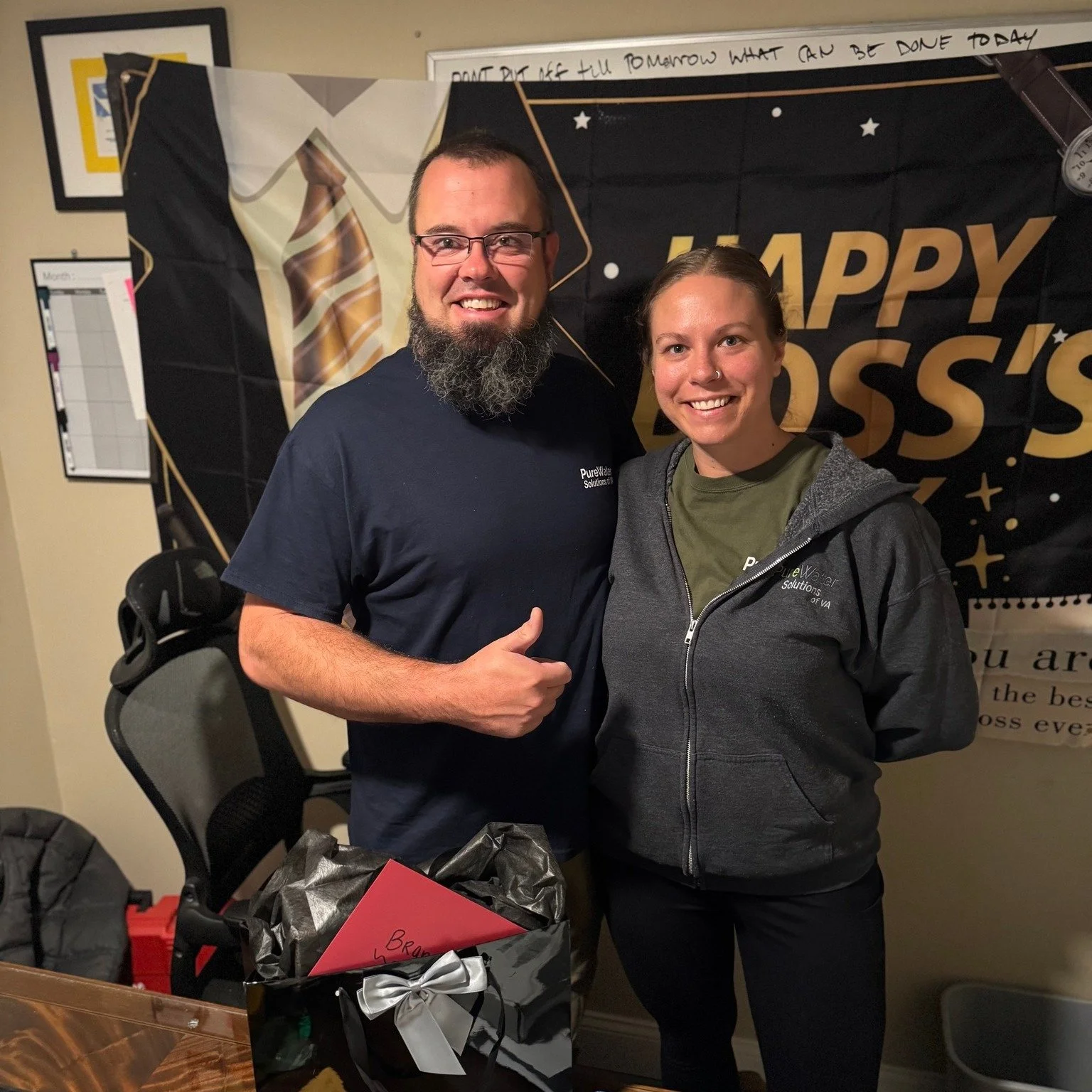 A man and woman smiling, standing together at an event with a black and gold banner behind them reading 'Happy Boss's Day.' The man has glasses and a beard, wearing a navy shirt. The woman has a nose piercing, wearing a gray hoodie and green shirt. There is a gift box with a silver ribbon on the table in front of them.