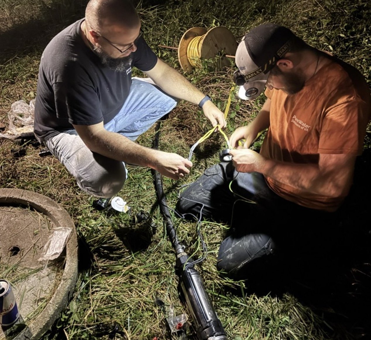 Two men working together outdoors at night, repairing or maintaining equipment involving a long black cylindrical device and various tools, with grassy ground and a plastic spool nearby.