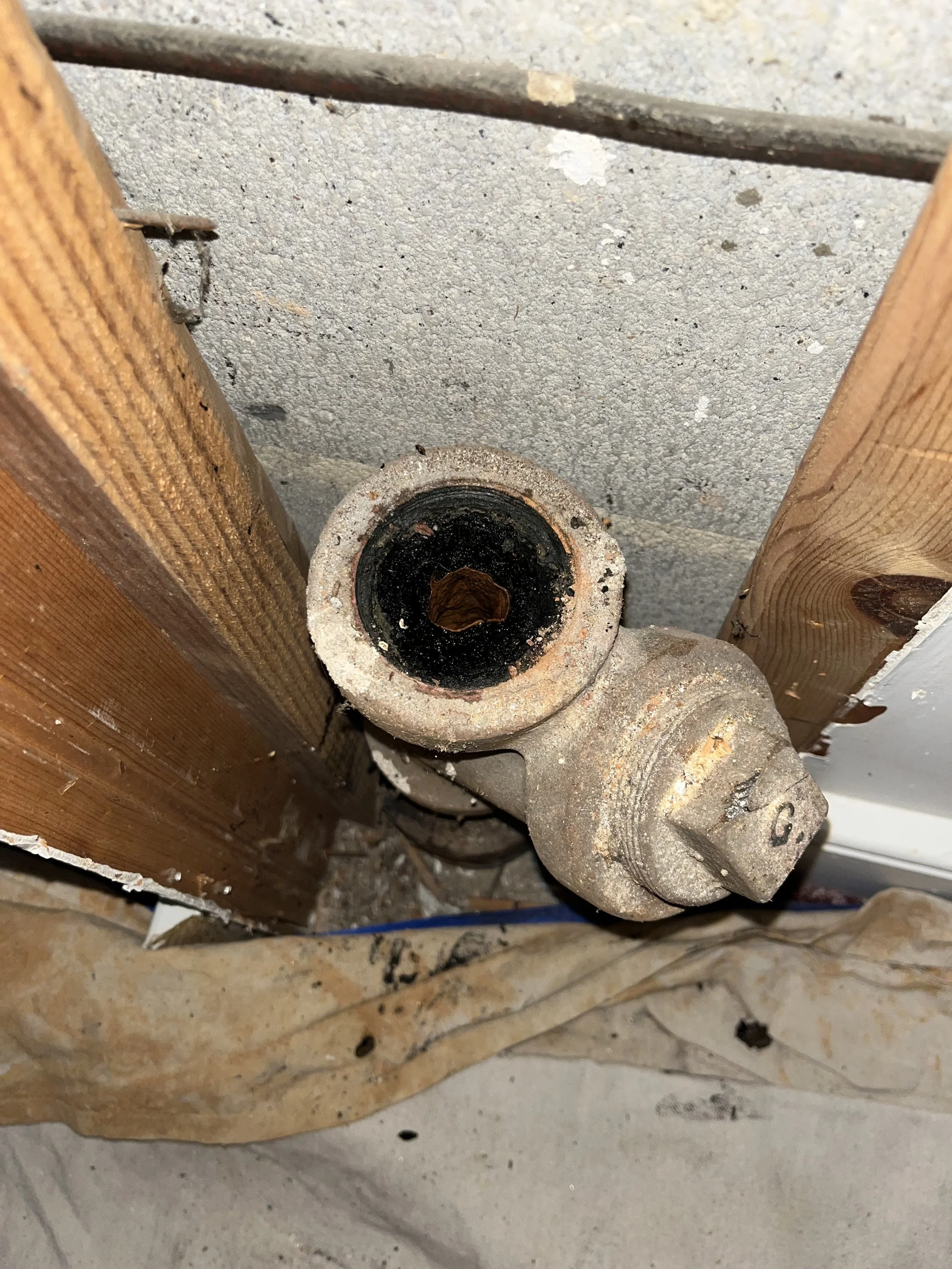 Close-up of a corroded pipe fitting or valve connection between a wooden post and a concrete wall, with black residue inside.