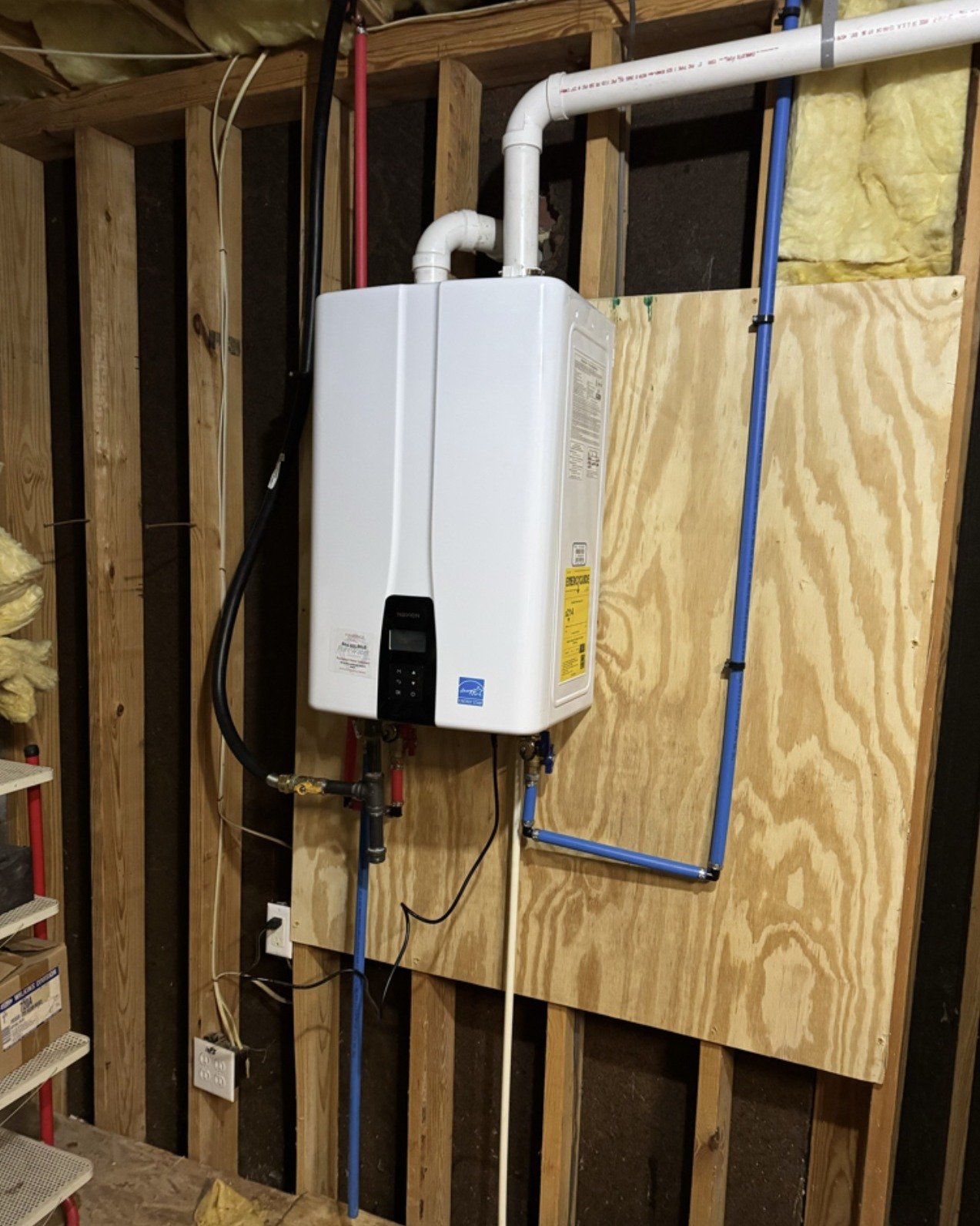 A residential tankless water heater installed on a plywood board in an unfinished basement with exposed wood framing, electrical wiring, and plumbing pipes.