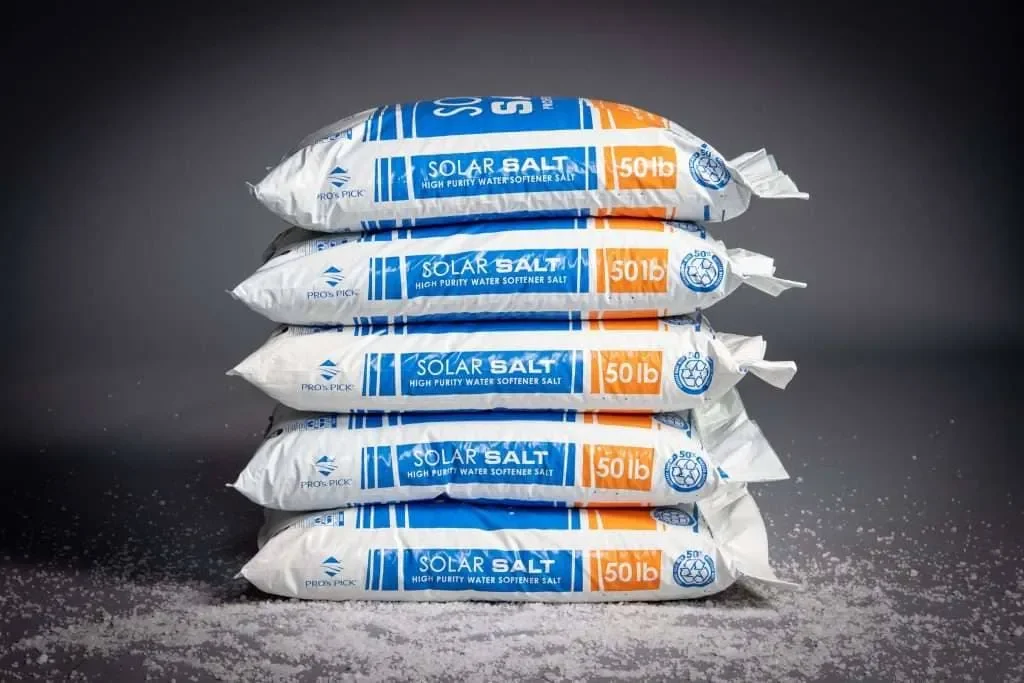 Stack of six 50-pound bags of solar salt, with some salt spilled on black surface.