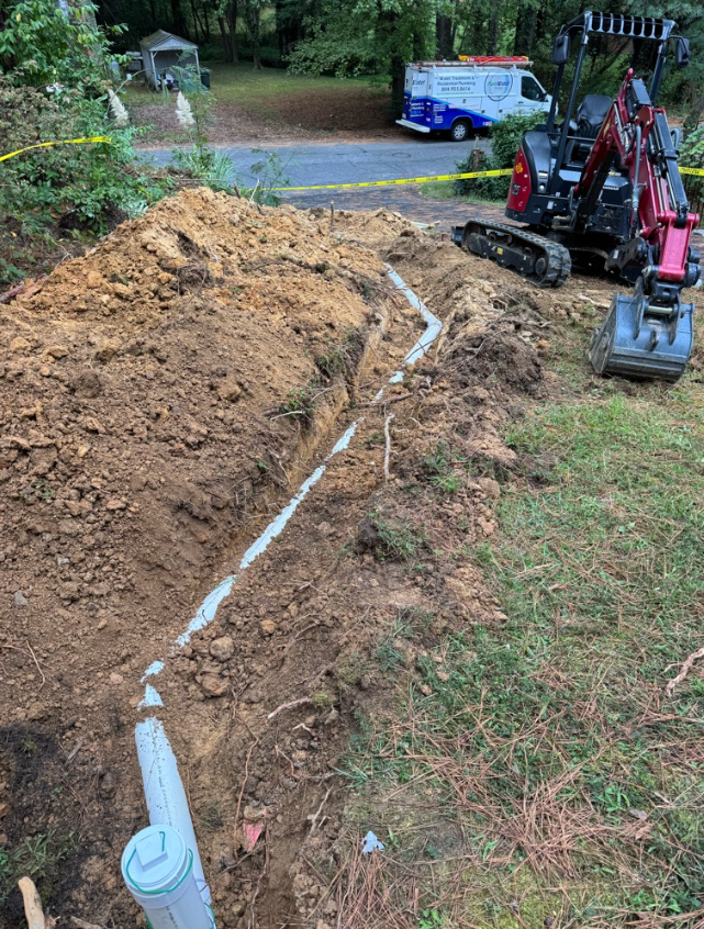 Excavation site with a trench dug for installing underground pipes, a backhoe, and a utility truck nearby, surrounded by trees and grass.