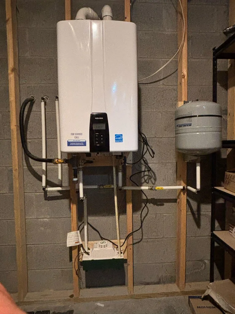 An electric tankless water heater installed on a wooden frame against a concrete wall, with pipes and electrical connections.