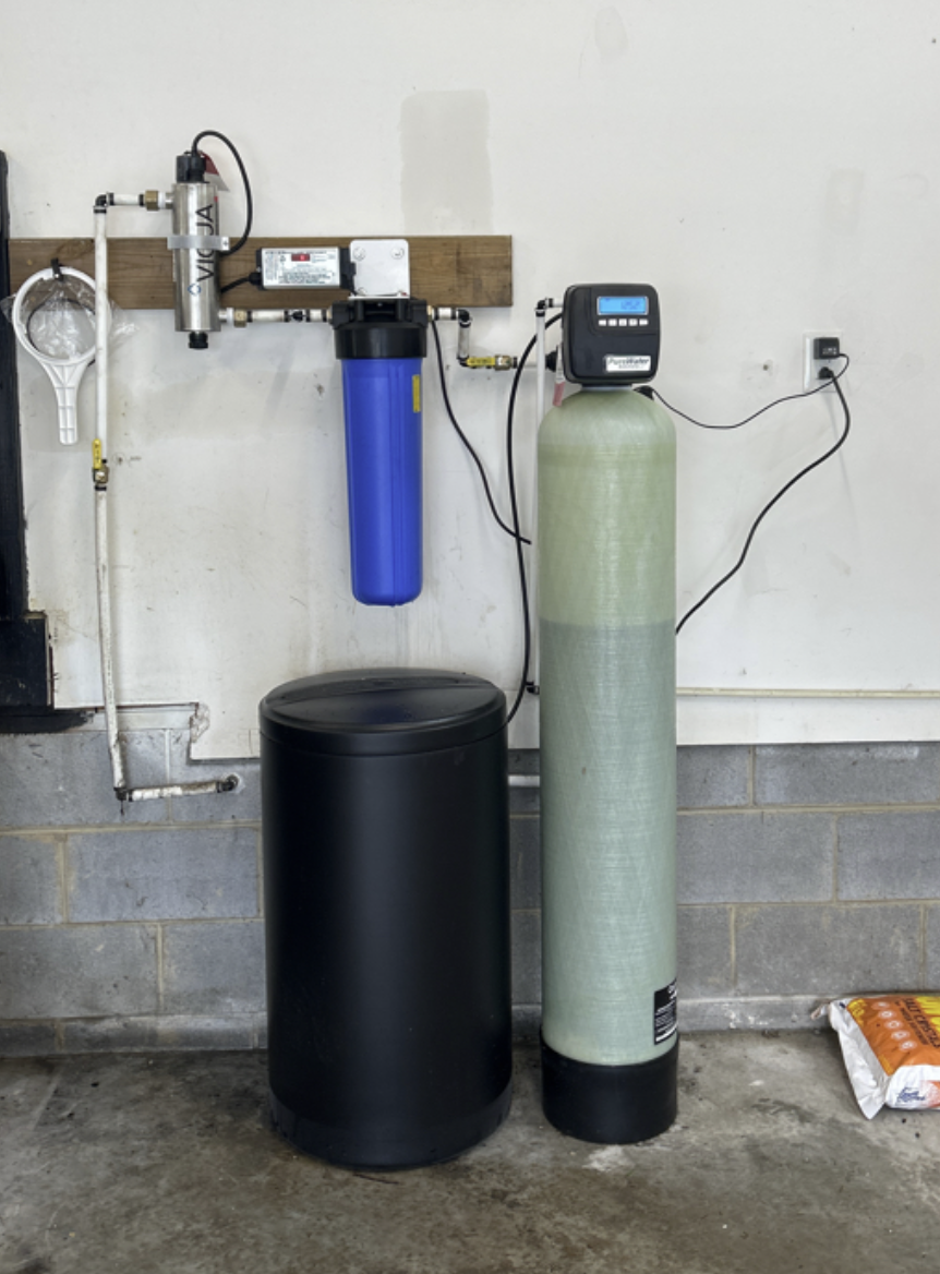 Water filtration system with multiple components including a blue filter housing, a green pressure tank with a control head, and pipes connected to a wall in a basement or utility room.
