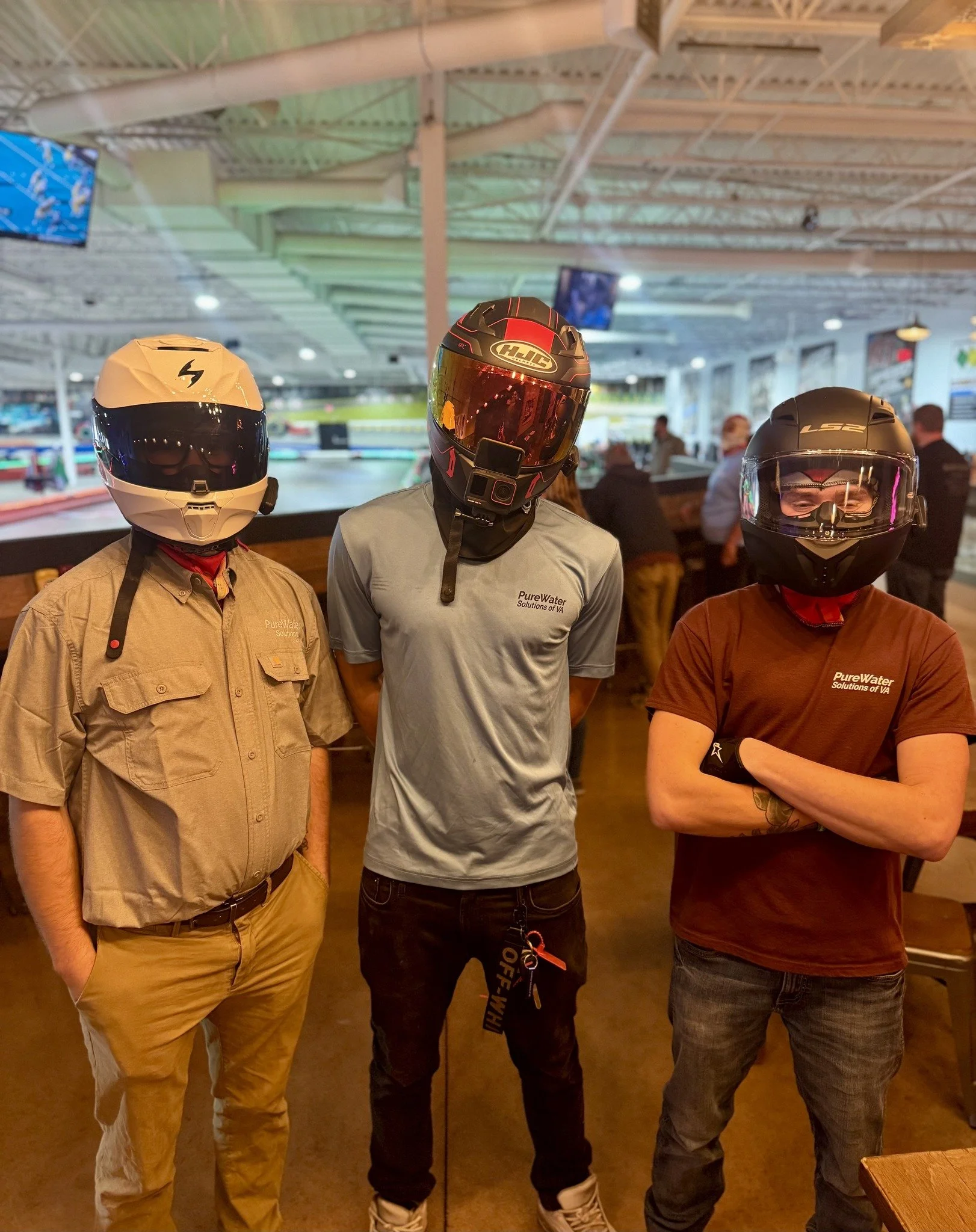 Three men wearing motorcycle helmets and casual clothes standing together indoors, with a racing track in the background.