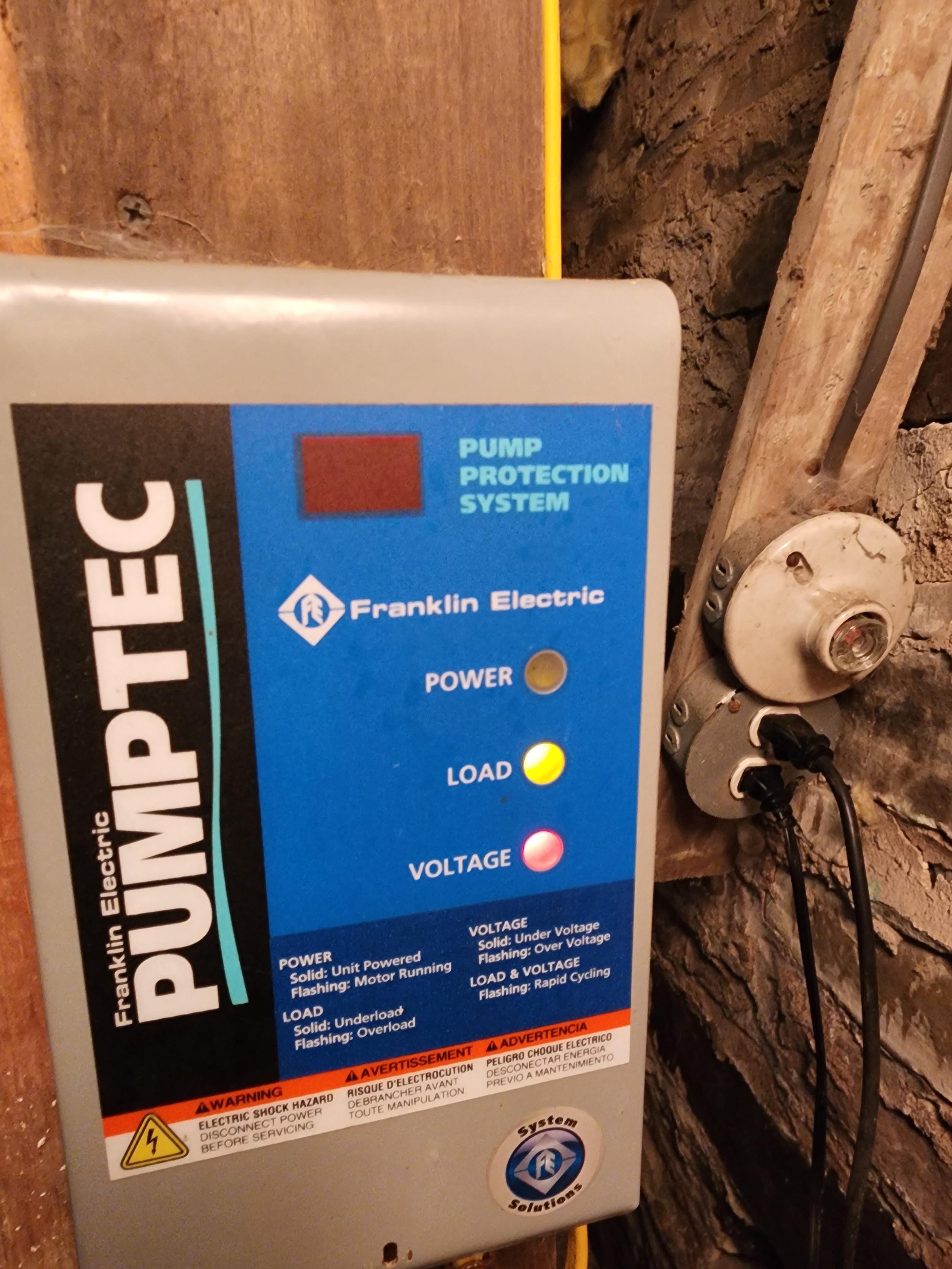 Electrical pump protection system control panel with indicator lights labeled power, load, and voltage, mounted on a brick wall with connected wires.