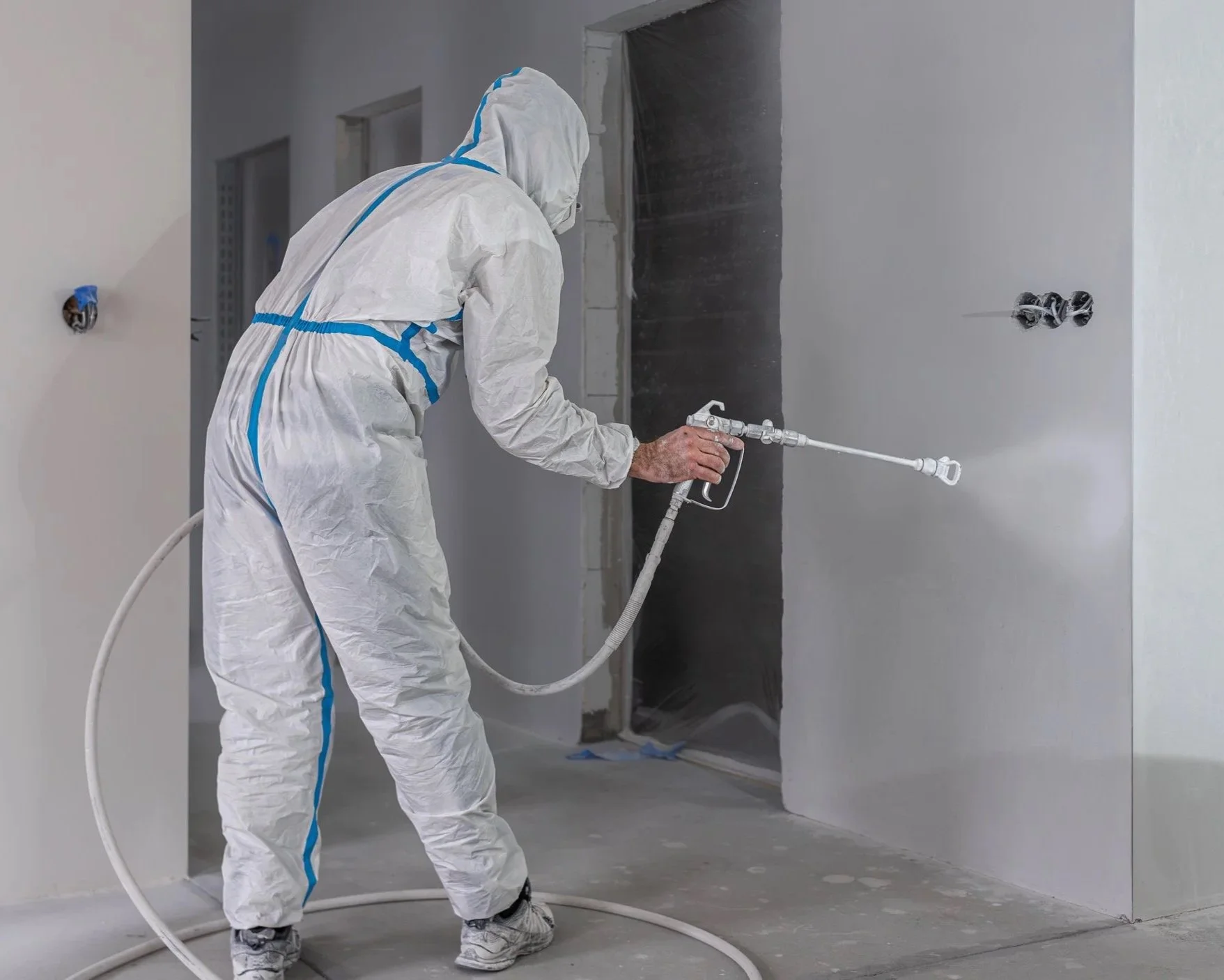 A worker wearing a full protective suit and mask is spraying a white wall with a spray gun in an indoor construction or renovation setting.