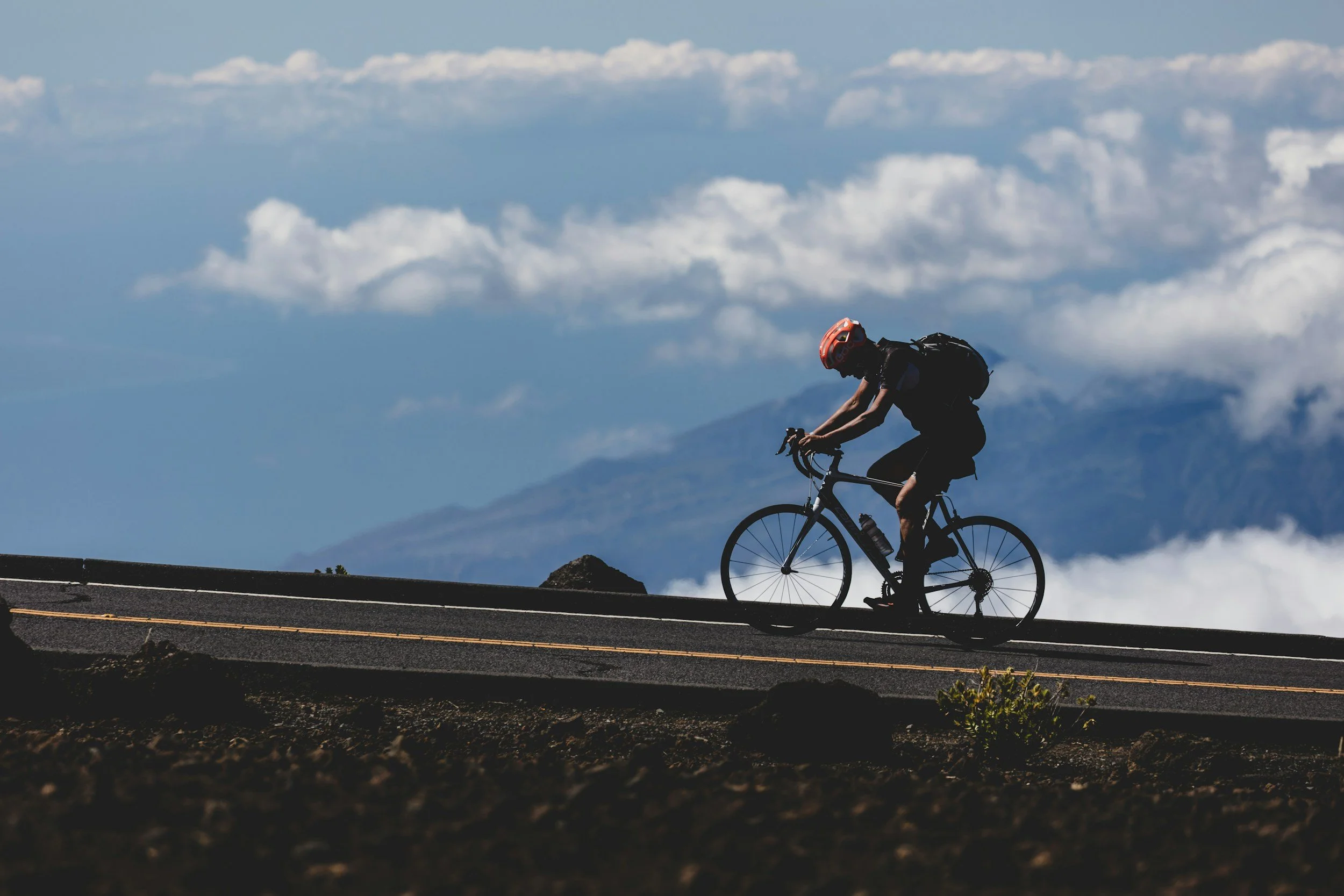 Professional cyclist riding mountain bike on mountain road with clouds and mountains in the background.